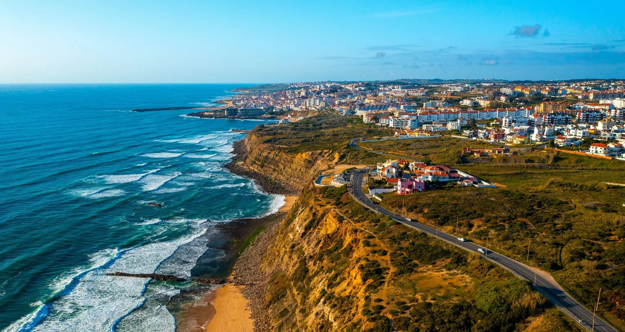 Aerial view of coastal city with cliffs, sandy beach, ocean waves, and numerous buildings under a blue sky.