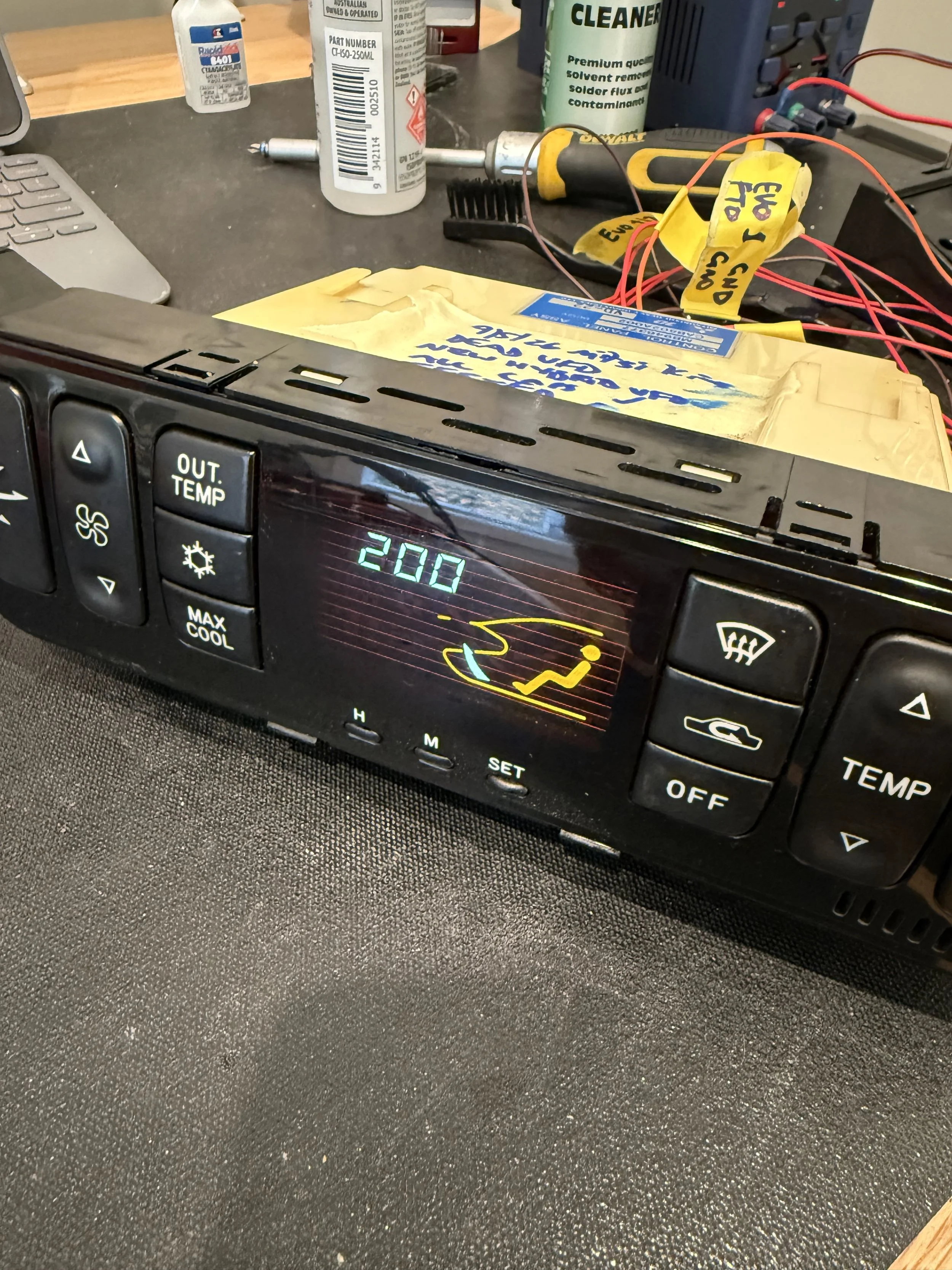 Evo car climate control panel displaying 20.0 degrees Celsius, with buttons for temperature adjustment, fan speed, and defrosting. In the background, various tools, wires, and cleaning supplies are scattered across a workbench.