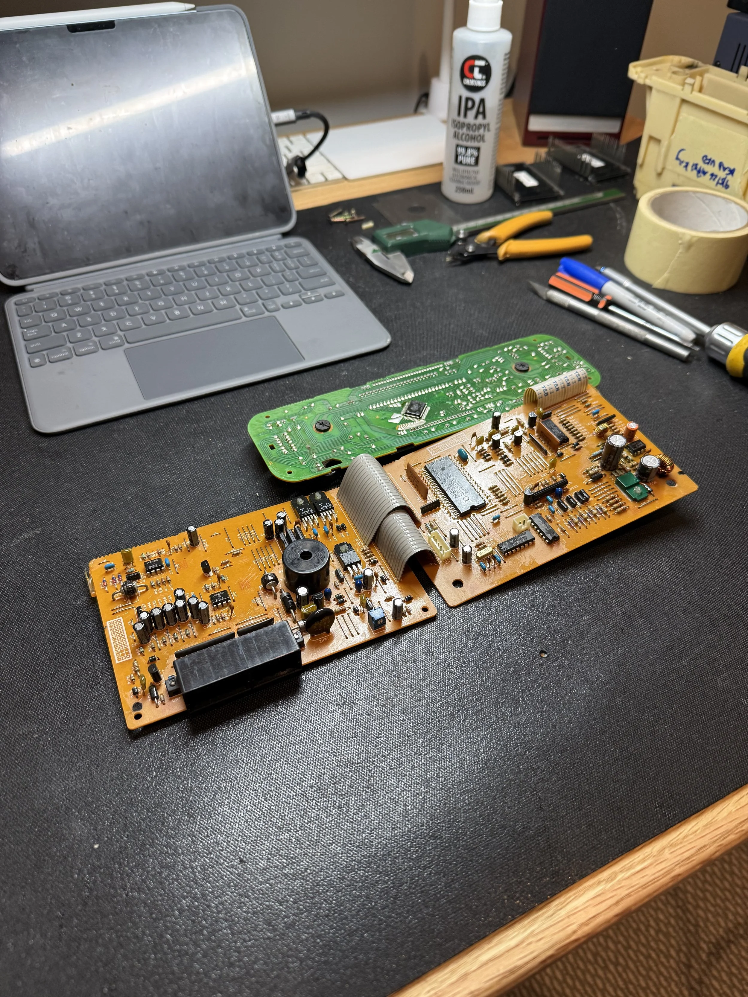 A workbench with electronic circuit boards, a laptop, tools, a bottle of IPA (isopropyl alcohol), a roll of masking tape, and other small components and tools.