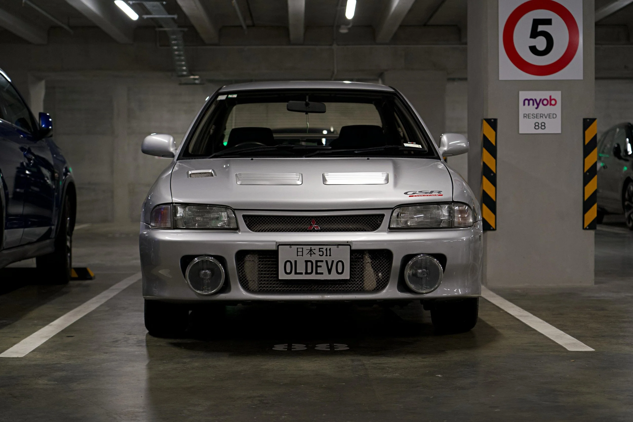 A silver Mitsubishi car with a Japanese license plate reading "OLDEVO" parked in an indoor parking garage. The front of the car faces the camera, and there are parked cars on either side. A speed limit sign of 5 km/h and a reserved parking sign with the number 88 are visible on a pillar behind the car.