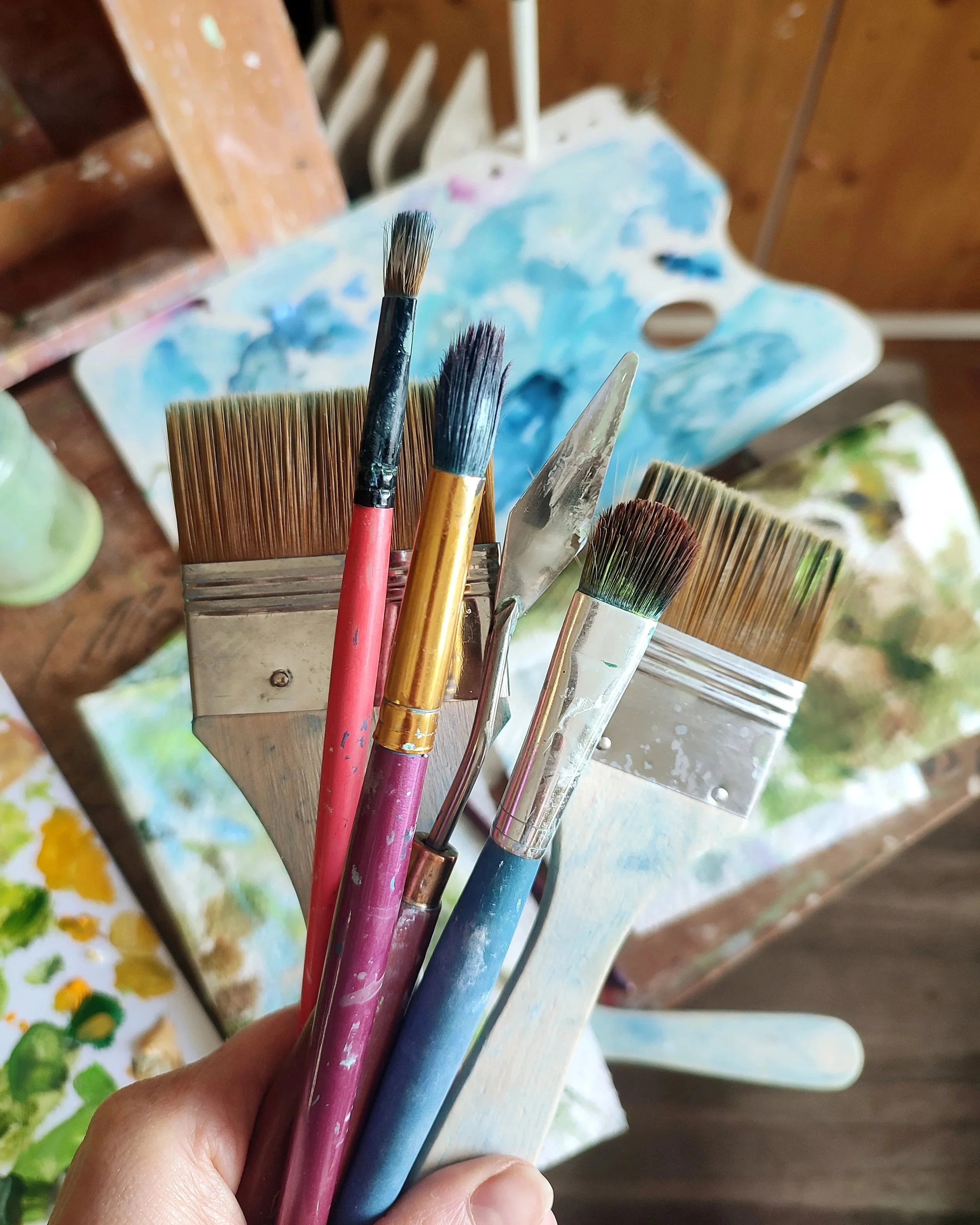 Close-up view of various paintbrushes held in a hand, with an artist's palette and a canvas in the background.