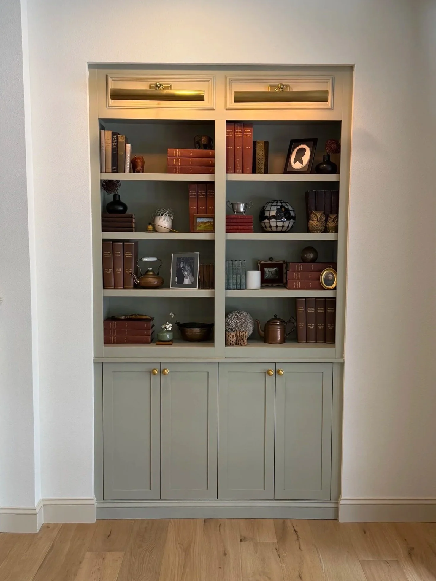 A bookshelf with four shelves filled with books, decorative objects, and framed photos, enclosed in a light gray cabinet with gold knobs, against a plain wall and wooden floor.