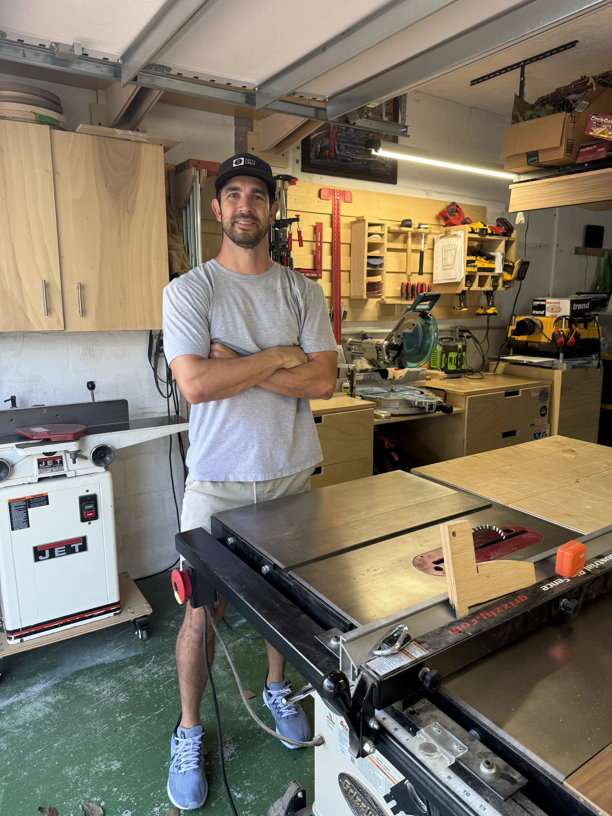 Man standing in woodworking workshop, crossing arms, wearing gray t-shirt, shorts, gray sneakers, and black cap, surrounded by woodworking tools and equipment.