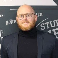 Un homme avec des lunettes, barbe rousse et porte un manteau noir, posant devant un mur avec des écritures en français