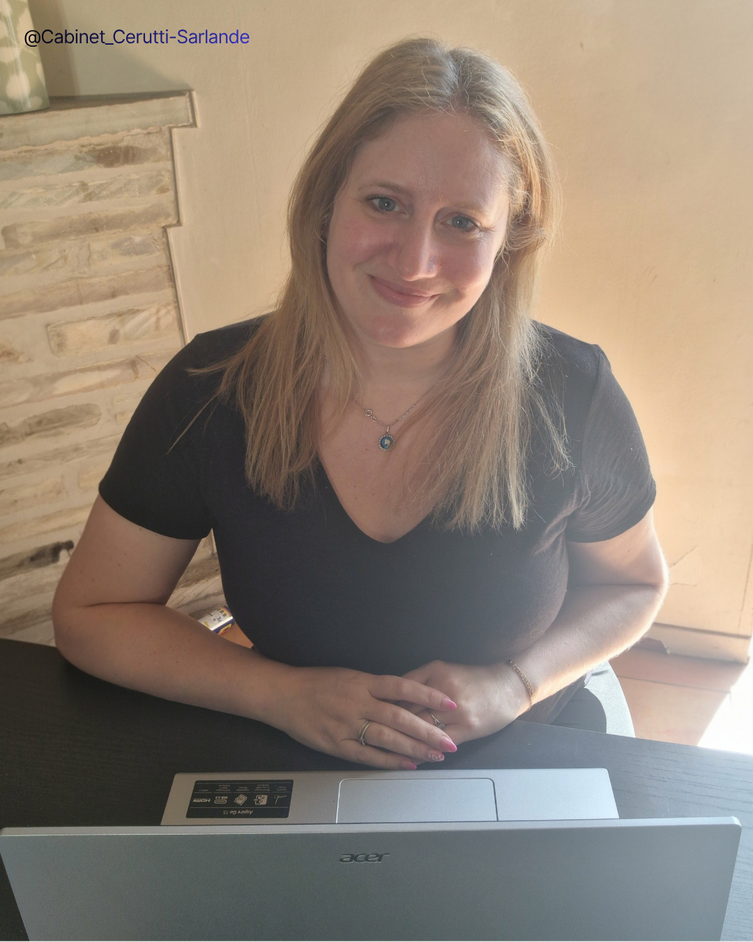 Femme assise à un bureau avec un ordinateur portable, souriante, portant un collier avec un pendentif, vêtue d'un t-shirt noir, dans un intérieur lumineux.