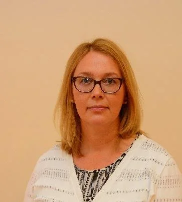 A woman with shoulder-length blonde hair, wearing glasses, a white cardigan, and a patterned top, standing against a beige wall.