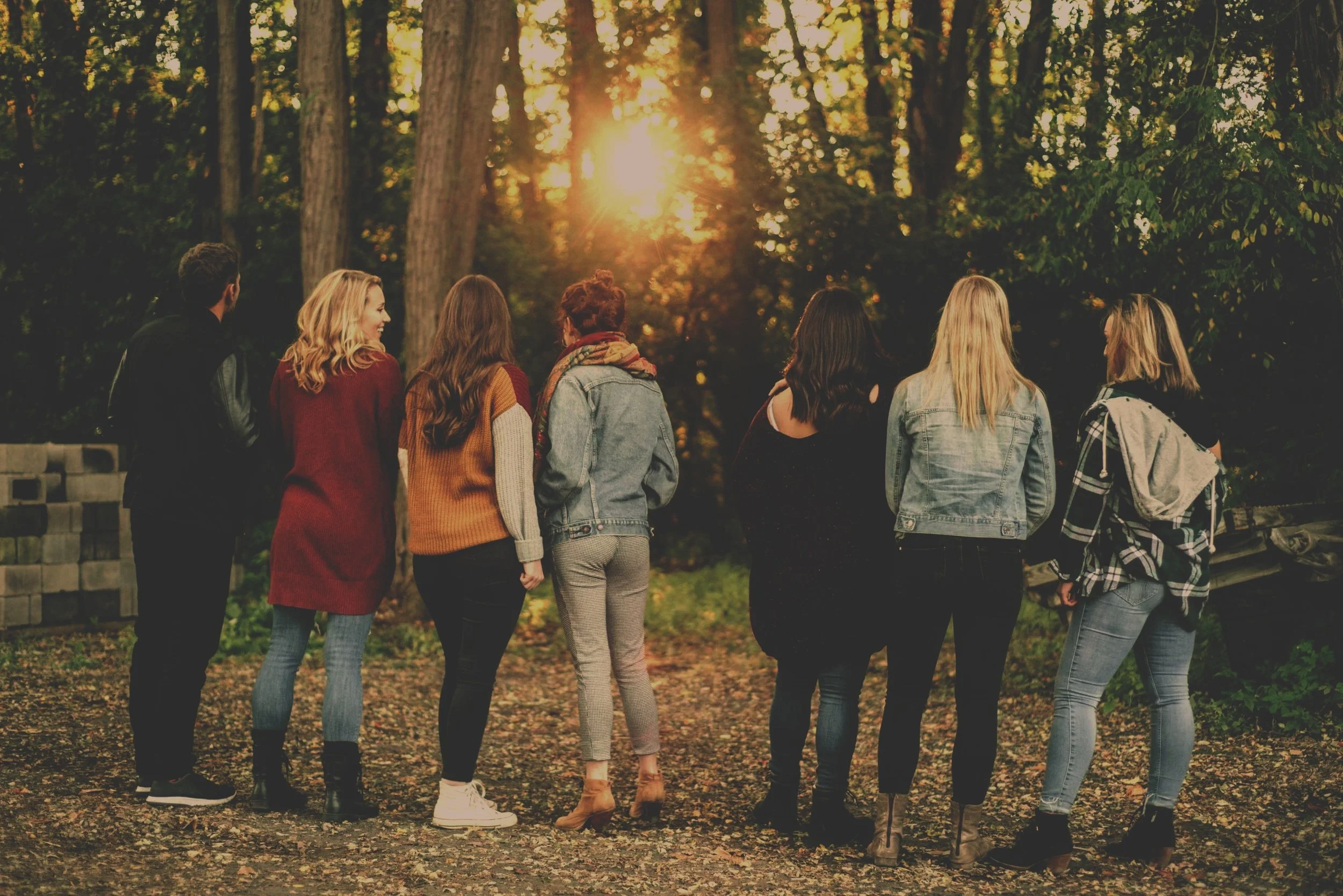 A group of eight people standing outdoors in a wooded area during sunset, facing away from the camera.