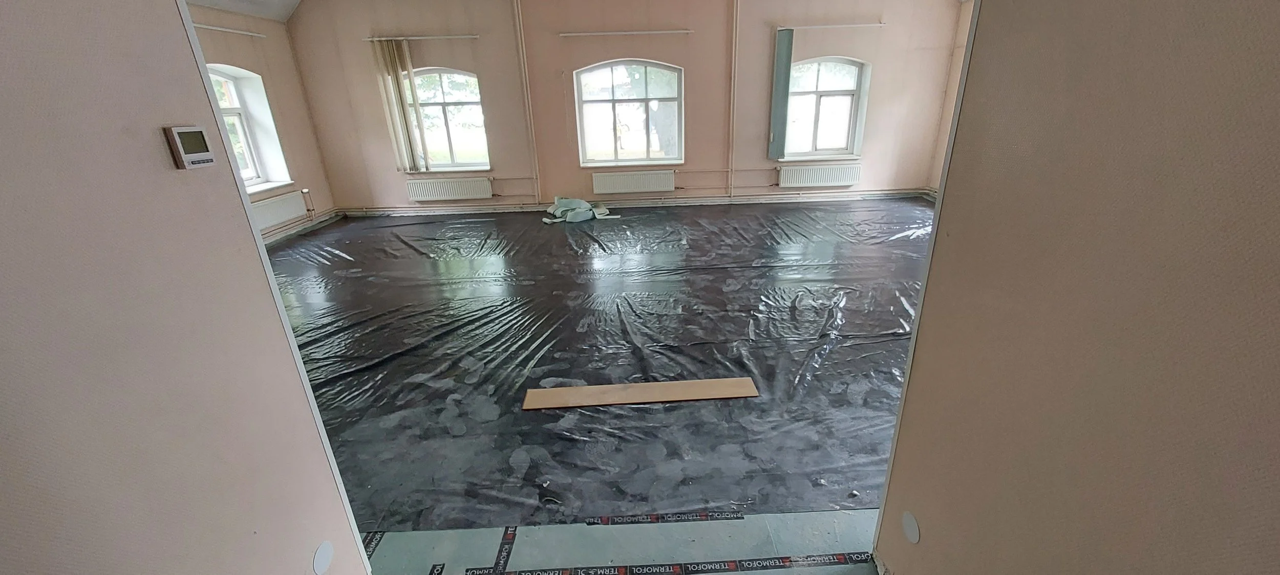 Empty room with three windows, radiator heaters, light pink walls, black plastic covering the floor, and some construction materials on the floor.