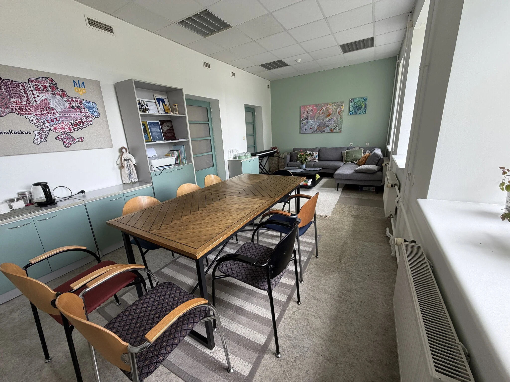 A meeting or conference room with a wooden table, chairs, a sofa with cushions, artwork on the walls, and a bookshelf with books and framed documents.