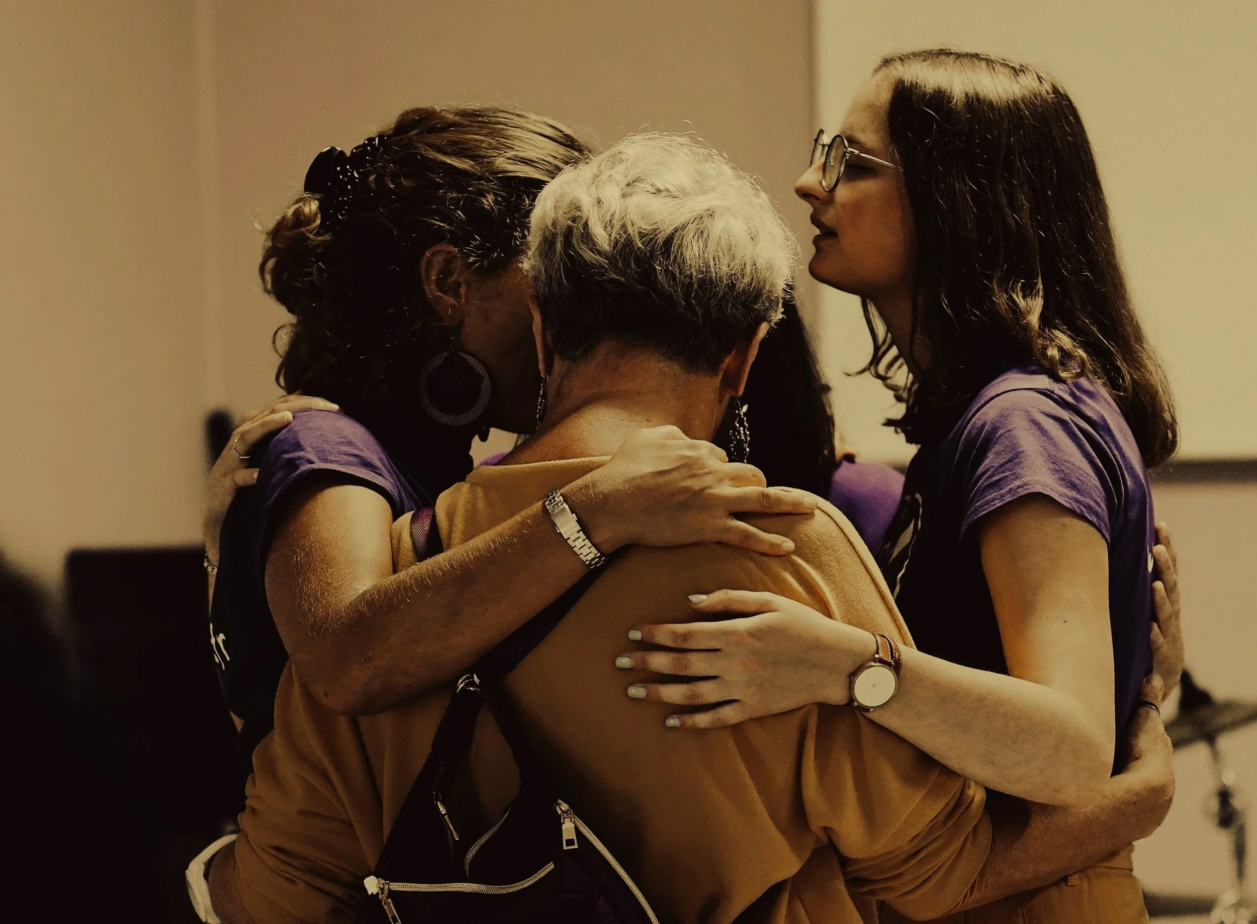 Four women hugging each other in a group embrace, standing close together indoors.