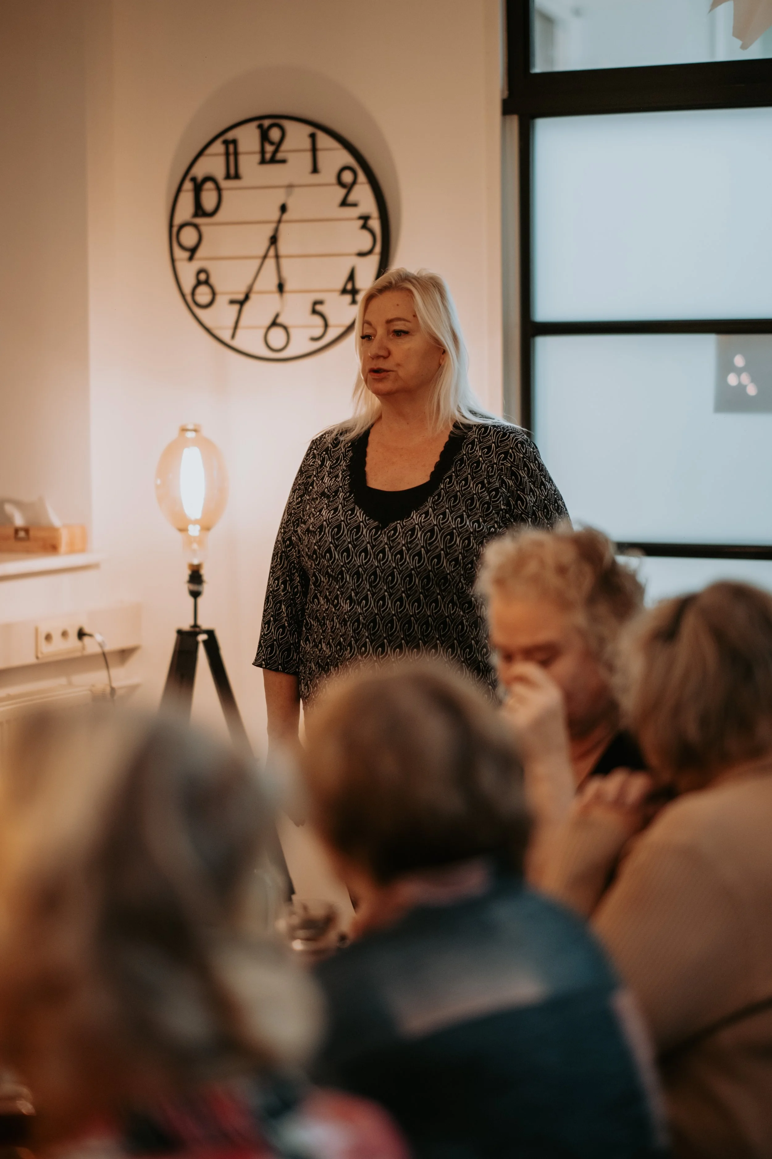 A woman with blonde hair standing in a room with a large wall clock and a window in the background, while a group of elderly people are seated and listening.