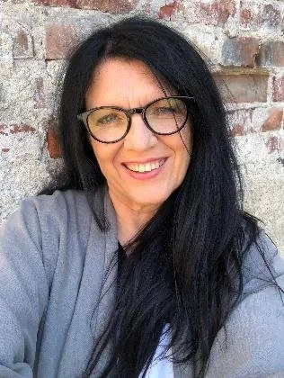 A woman with black hair wearing glasses and a gray top, smiling in front of a brick wall.