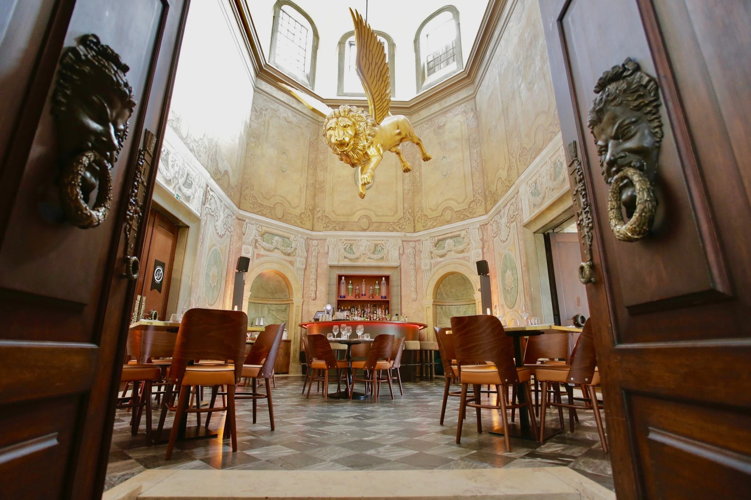 A film and photo location in Portugal from walnut films, with a golden winged lion sculpture hanging from the ceiling, ornate wall murals, wooden tables and chairs, and an elaborate doorway with lion head door knockers.