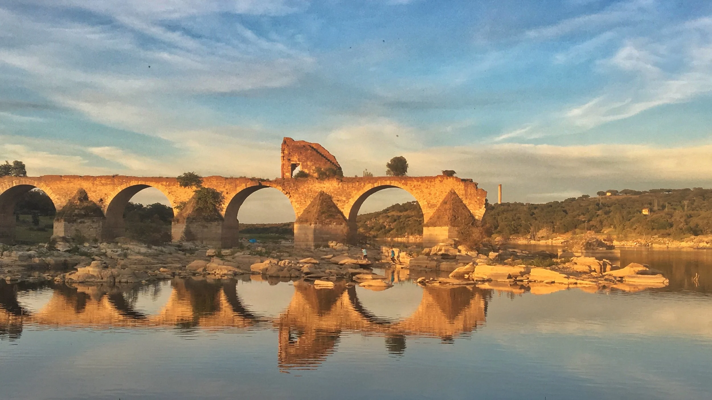 A film and photo location in Portugal from walnut films, Ancient stone aqueduct over a river with rocks on the riverbank, under a partly cloudy sky during sunset.