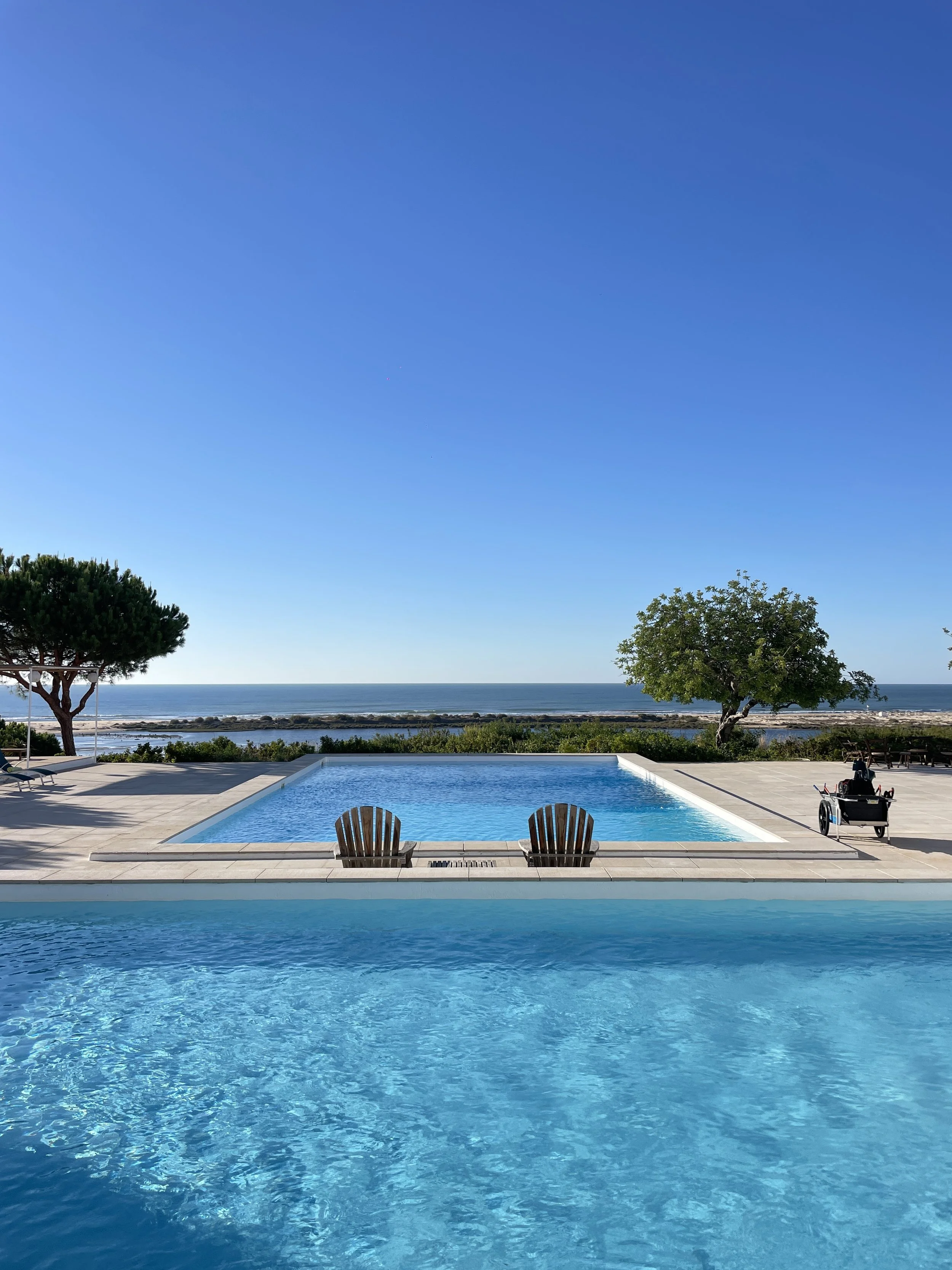 A film and photo location in Portugal from walnut films, Swimming pool with two wooden chairs facing the pool, overlooking the ocean with a clear blue sky, trees, and a distant shoreline.