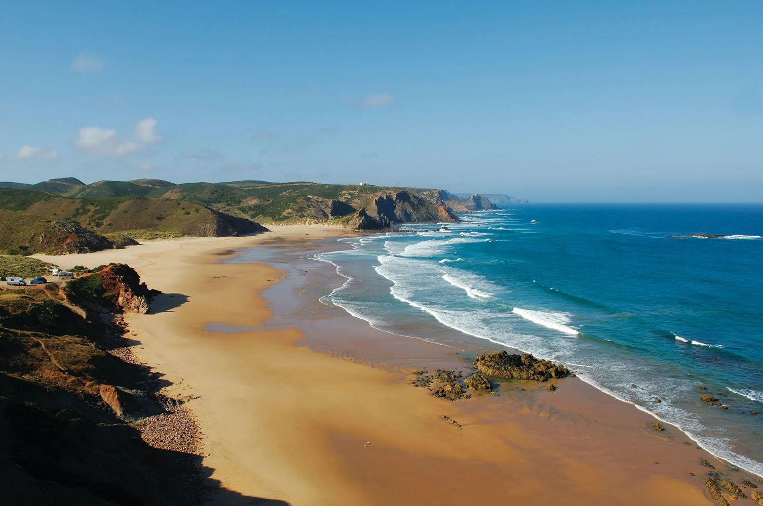 A film and photo location in Portugal, scenic view of a beach with golden sand, rugged cliffs covered in green vegetation, and the ocean with white waves under a clear blue sky.