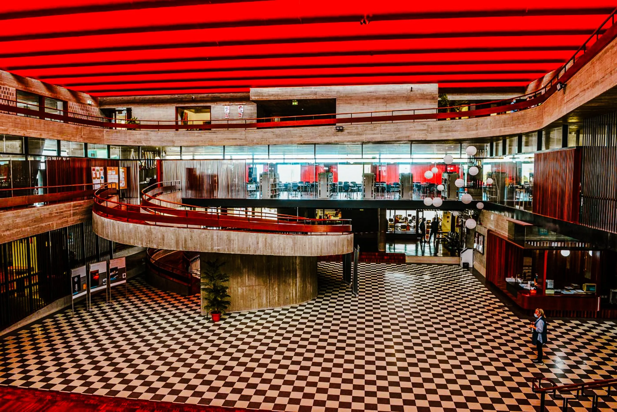 A film and photo location in Portugal from walnut films, Interior of a modern multi-story building with red ceiling accents, curved balconies with red railings, large windows, black and white checkered tile floors, and a few people walking.