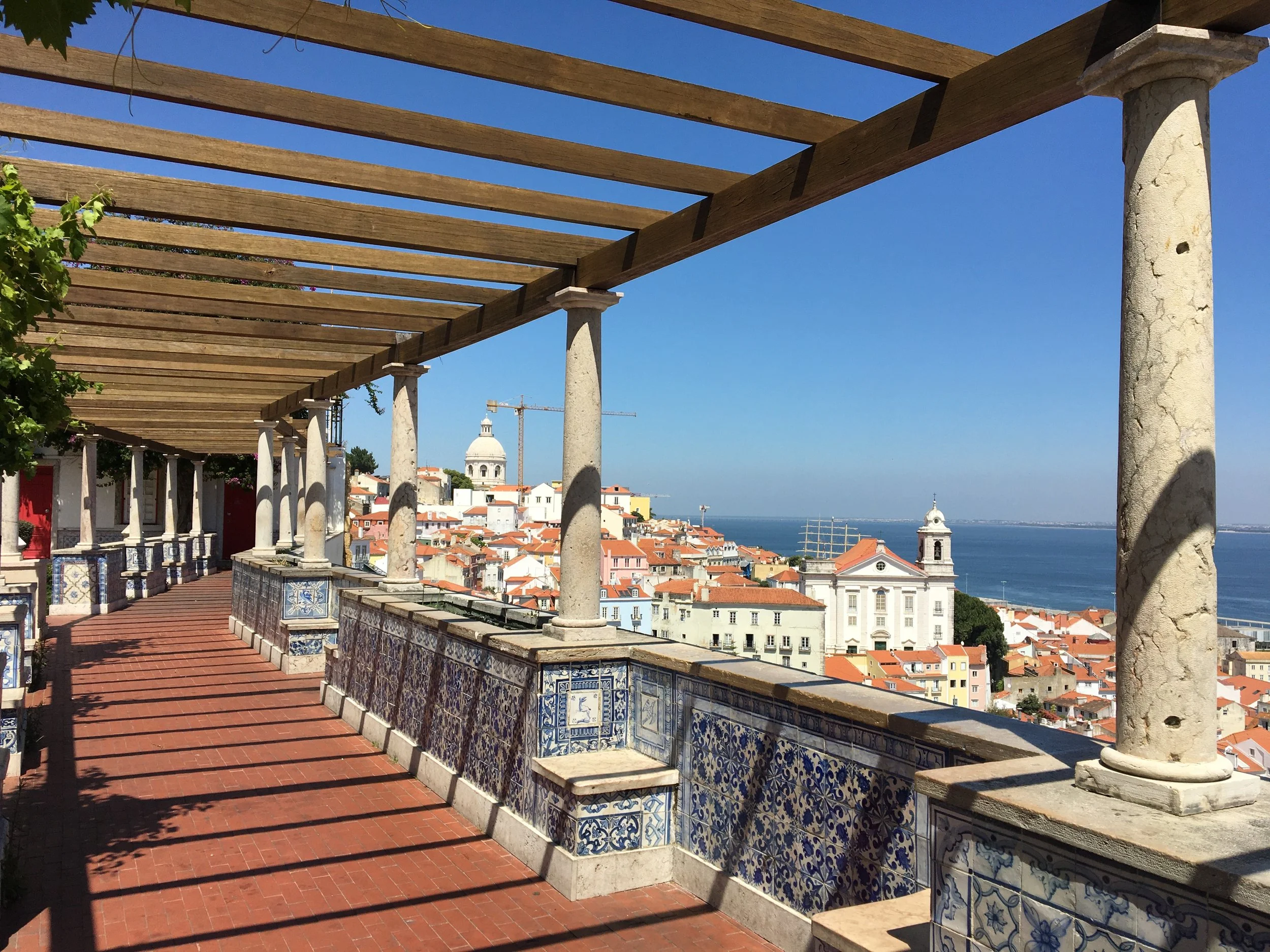 A film and photo location in Portugal from walnut films, View from a terrace with a wooden pergola, tiled railing, and overlooks a city with red-tiled roofs, church domes, and a body of water in the distance.