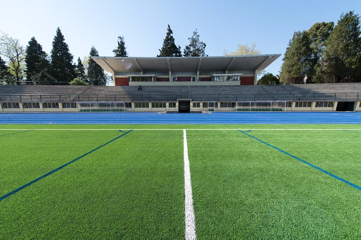 A film and photo location in Portugal from walnut films, Empty athletic stadium with a green field, blue track, and a modern stand with trees in the background under a clear blue sky.