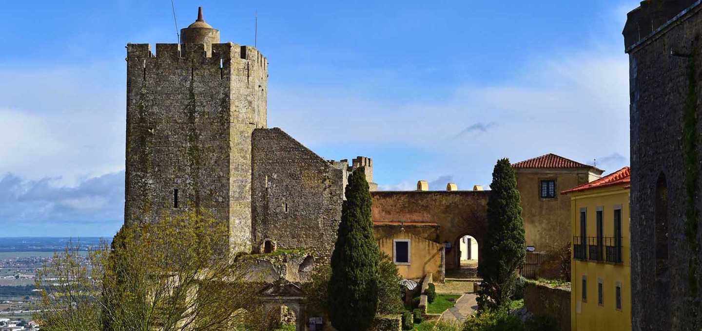 A film and photo location in Portugal from walnut films, Ancient stone castle with a tower and surrounding buildings, with trees and a cityscape in the background.