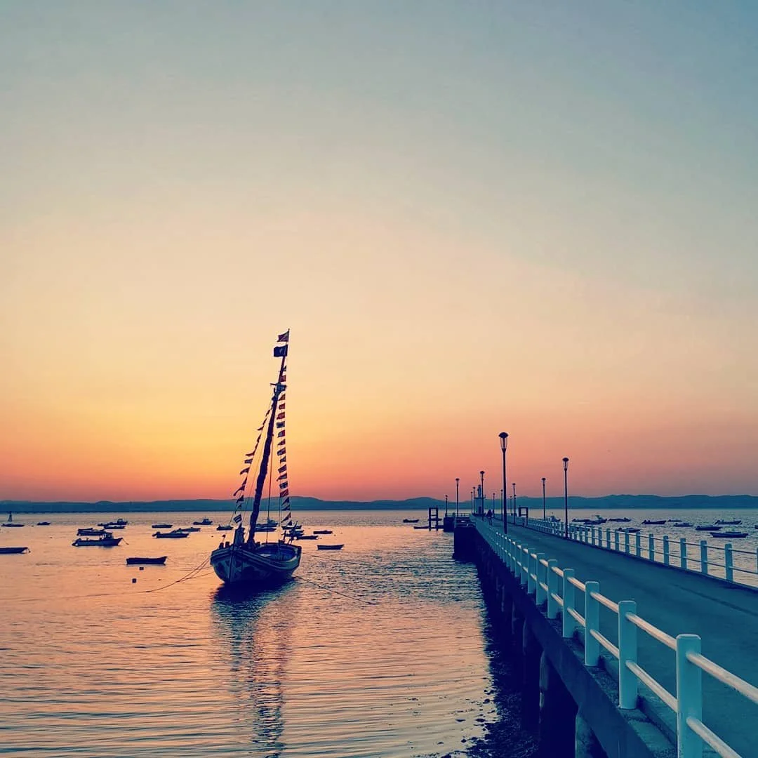 A film and photo location in Portugal from walnut films, A sailboat docked near a pier at sunset with smaller boats floating on the water and a colorful sky in the background.