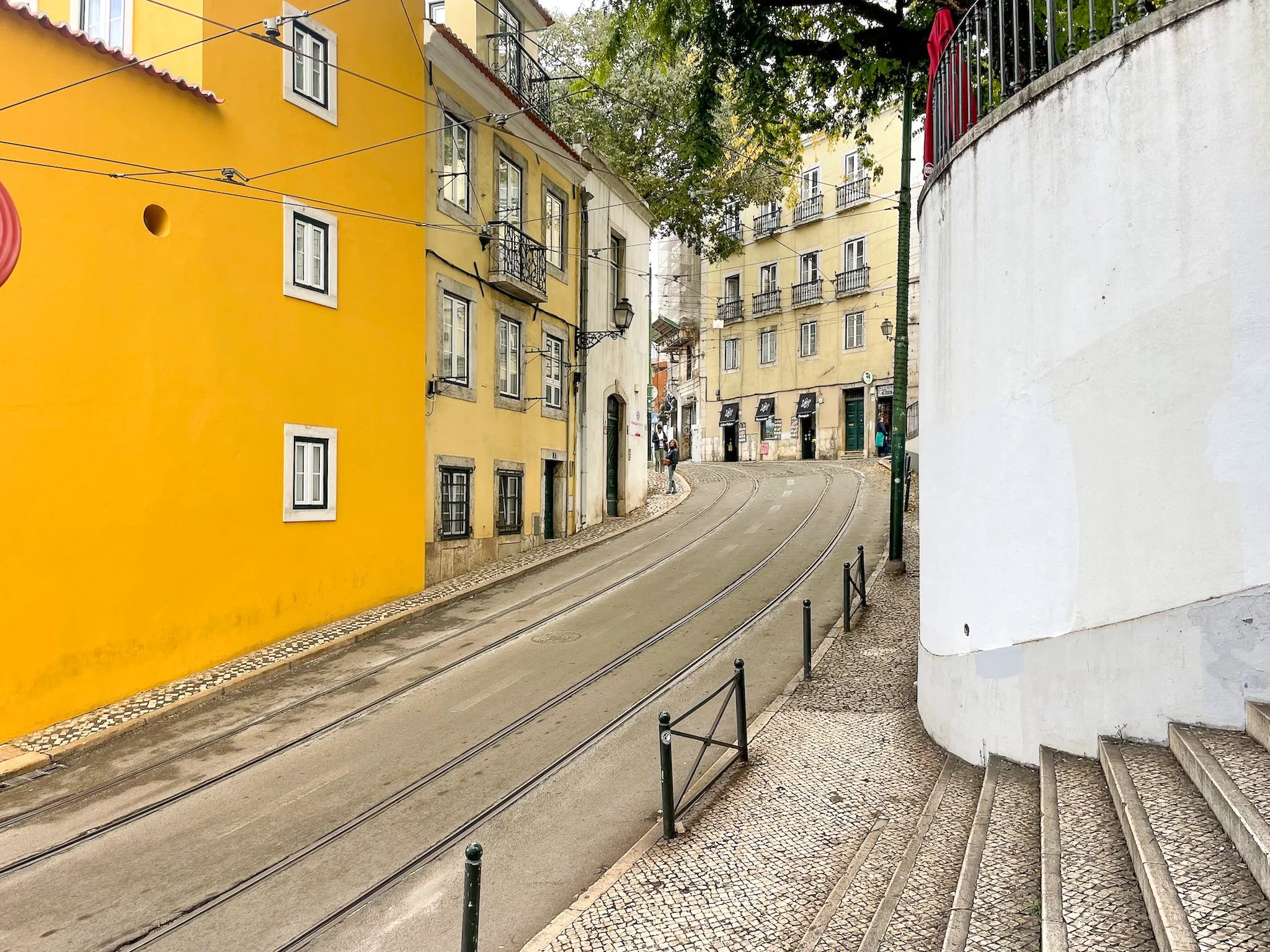 A film and photo location in Portugal from walnut films, A narrow street with tram tracks, surrounded by colorful buildings with yellow and white facades, cobblestone sidewalks, and a few people walking.