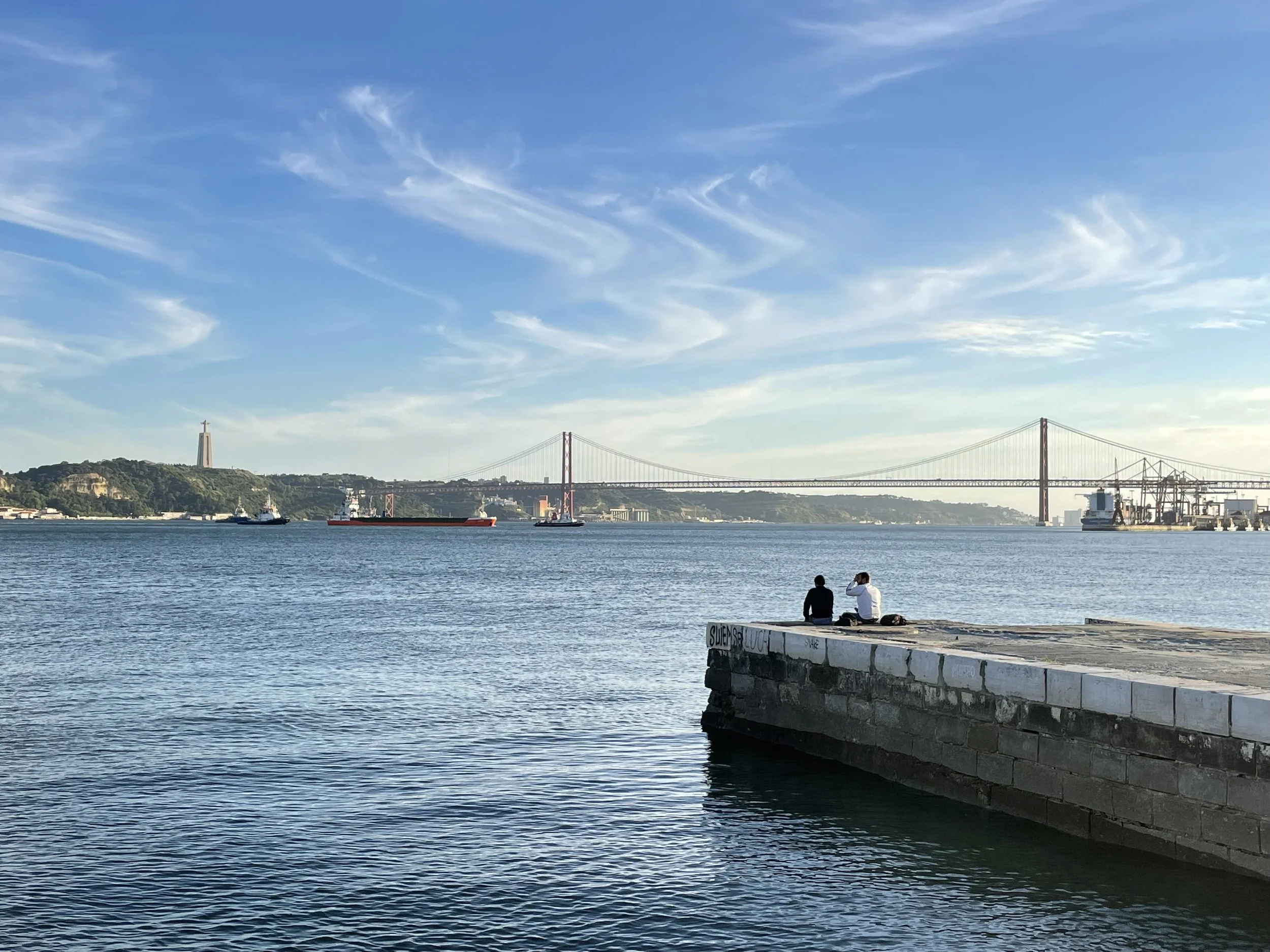 A film and photo location in Portugal from walnut films, View of a bridge spanning across a wide body of water, with a hilly landscape and a statue on top of a hill to the left. Two people sit on a pier in the foreground, 