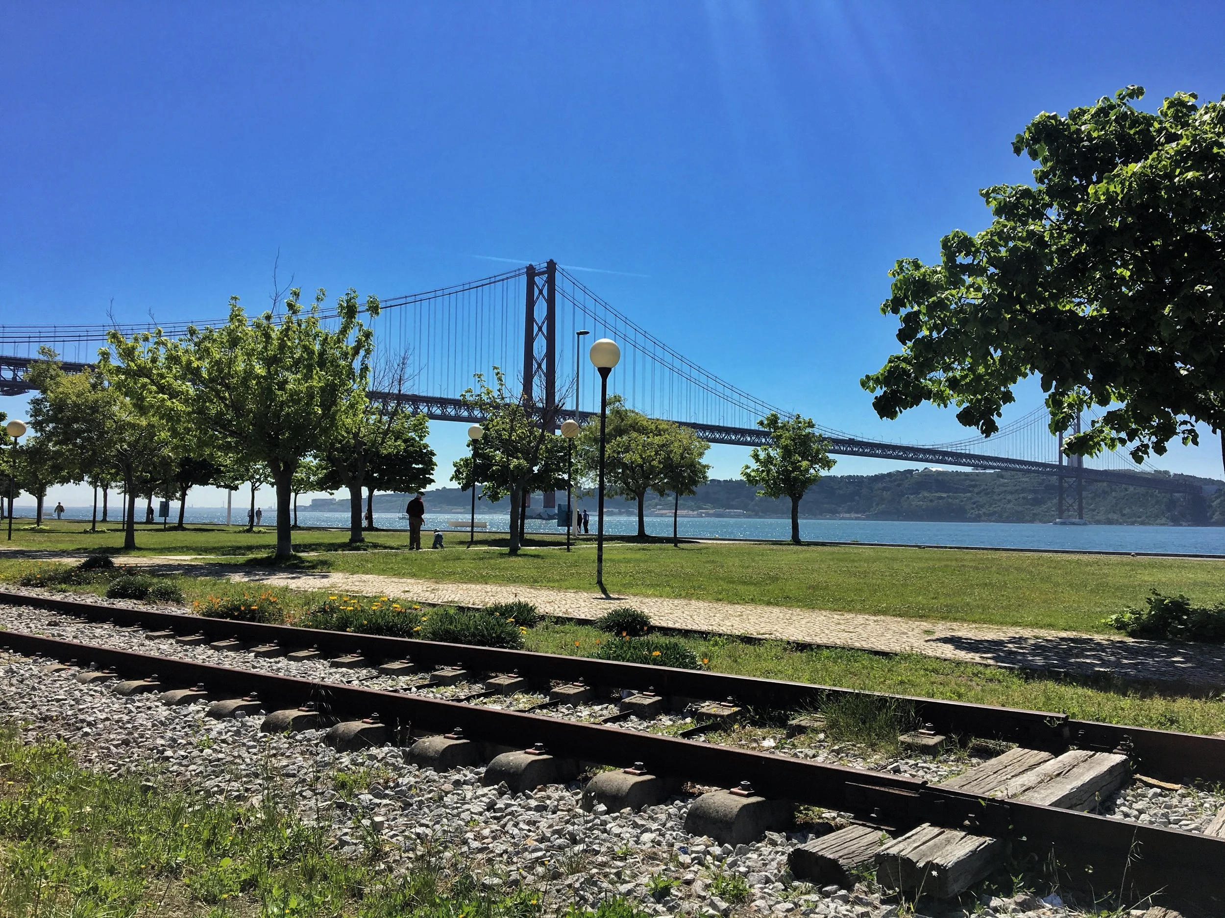 A film and photo location in Portugal from walnut films, A park with trees, benches, and street lamps along a river, with a bridge over the water and a clear blue sky.