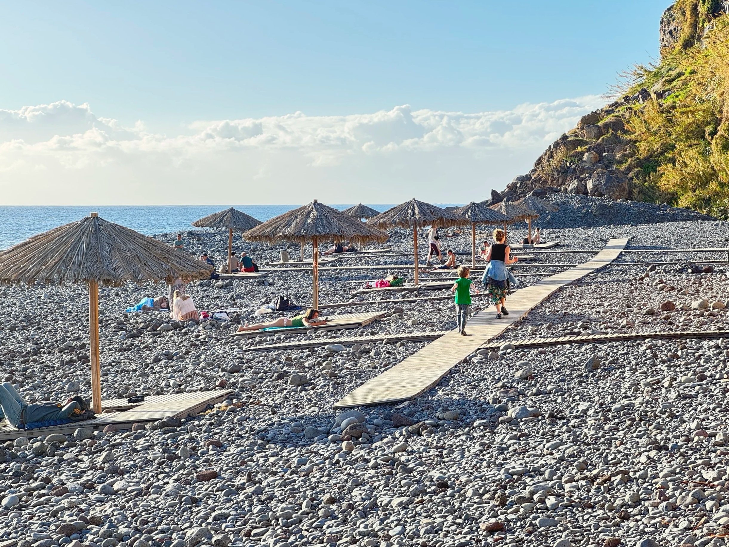 A film and photo location in Portugal from walnut films, A pebble beach with several straw umbrellas and people relaxing on lounge chairs or walking along a wooden pathway. A rocky cliff with green vegetation is on the right, and the ocean