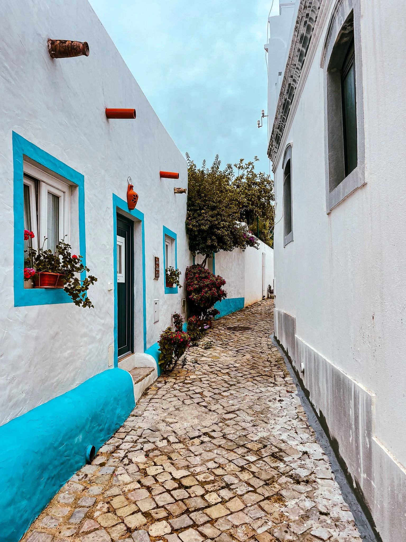 A film and photo location in Portugal from walnut films, A narrow cobblestone street between white buildings with colorful accents, potted plants, and flowers, under a blue sky.