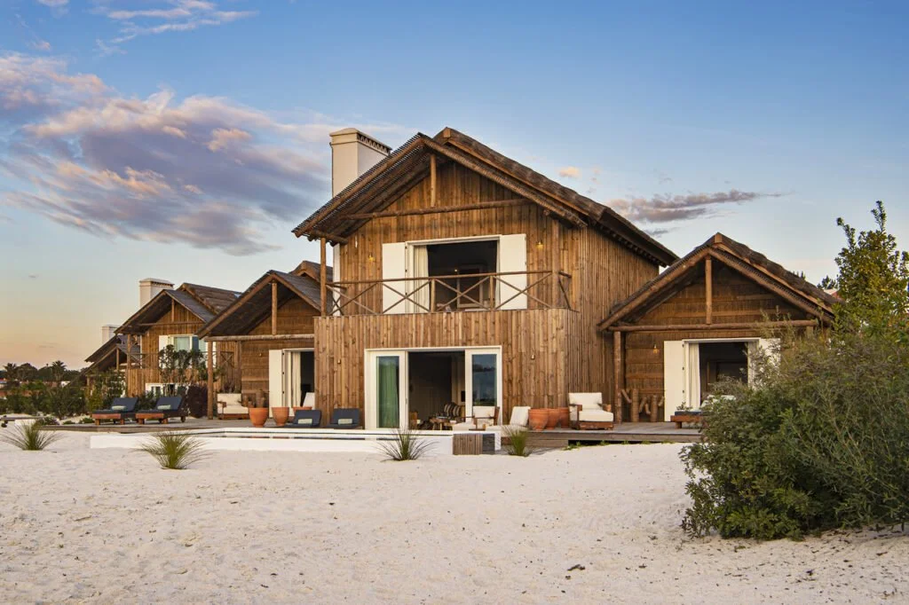 A film and photo location in Portugal from walnut films, A row of rustic wooden beach houses with outdoor seating on a sandy beach under a partly cloudy sky.