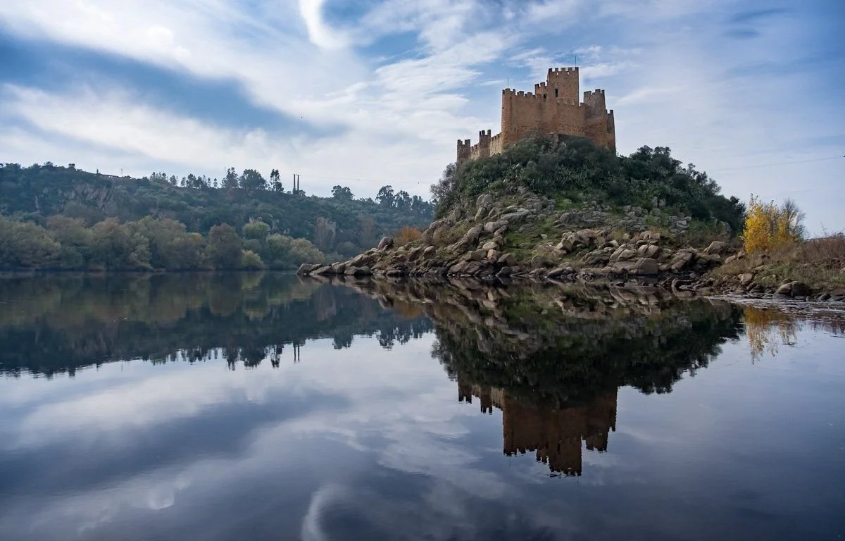 A film and photo location in Portugal from walnut films, A castle on a hill reflected in a calm body of water with a partly cloudy sky.