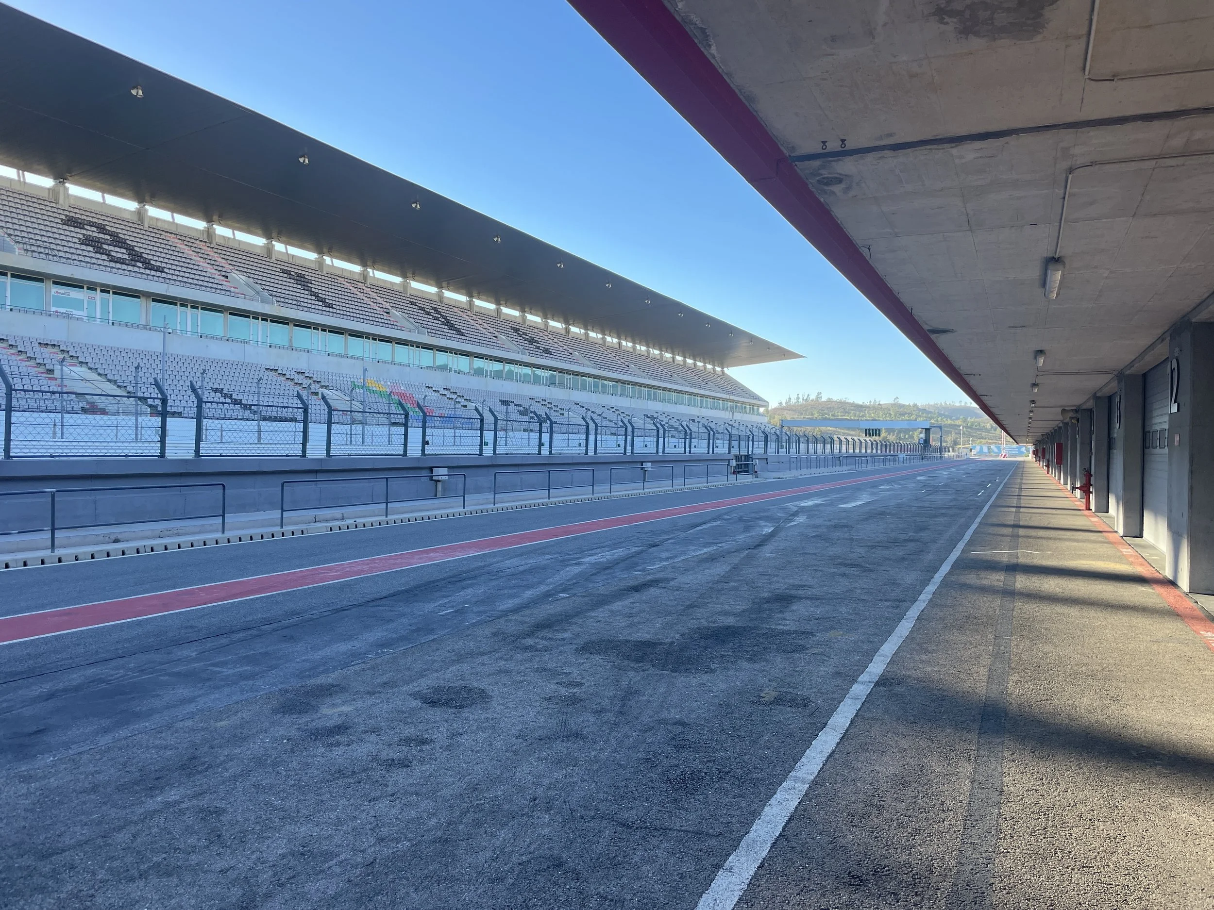 A film and photo location in Portugal from walnut films, View of an empty race track pit lane at a racing circuit, with grandstands and a hill in the distance under a clear blue sky.