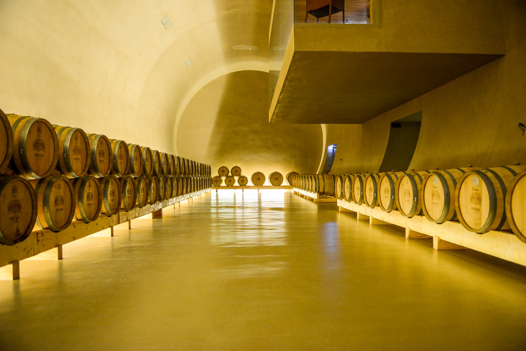 A film and photo location in Portugal from walnut films, A wine cellar room with rows of stacked barrels on wooden stands, with a yellowish wall and an arched ceiling, illuminated by warm lighting.