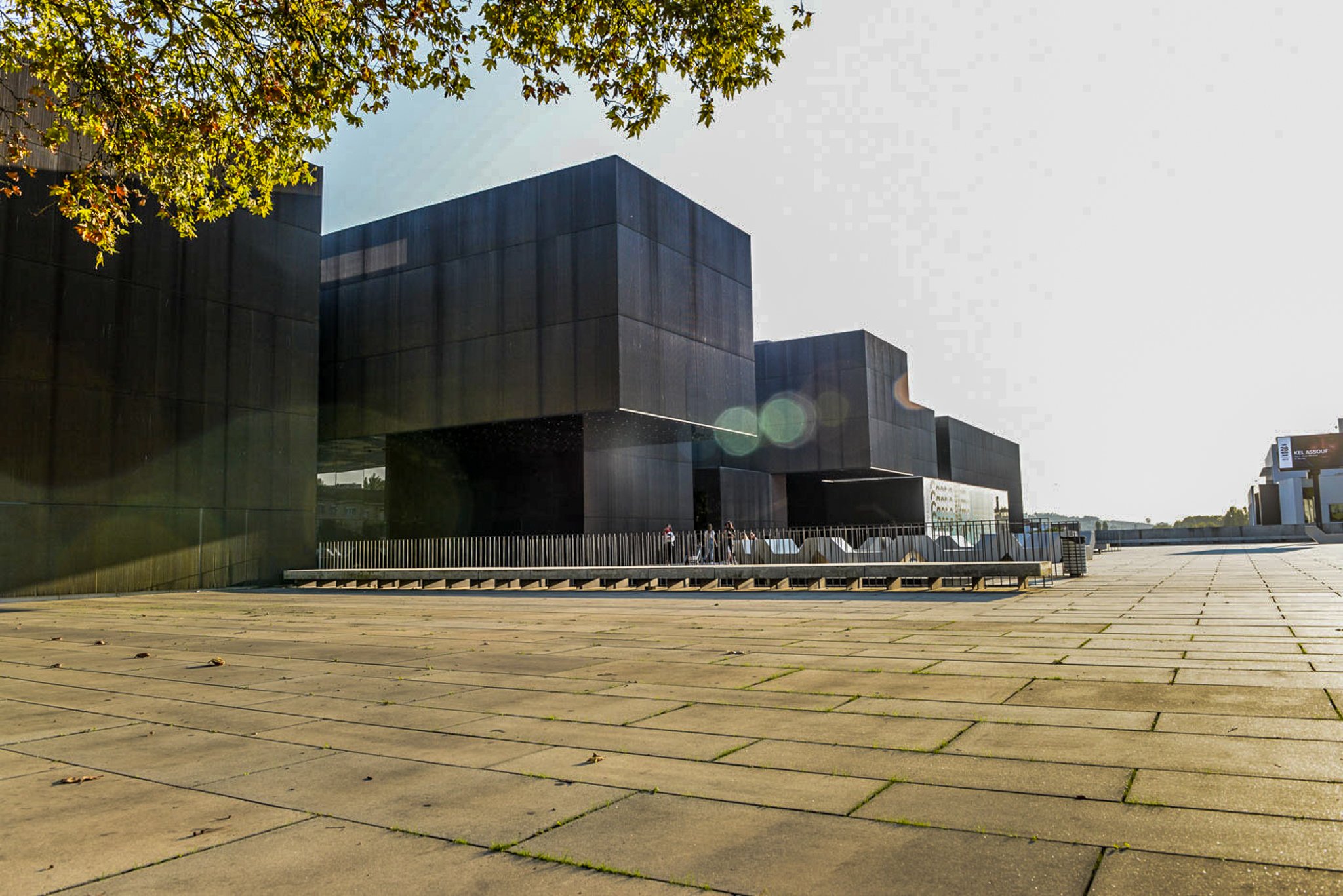 A film and photo location in Portugal from walnut films, Modern building with dark exterior panels, situated in an open plaza with wooden paving, under a clear sky with sunlight filtering through a tree with green leaves.