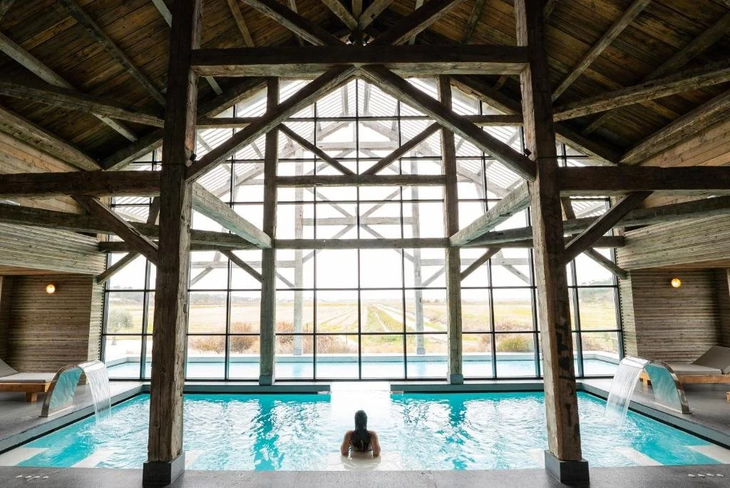 A film and photo location in Portugal from walnut films, Indoor swimming pool with a person swimming, surrounded by wooden beams and large glass windows exposing a countryside view.