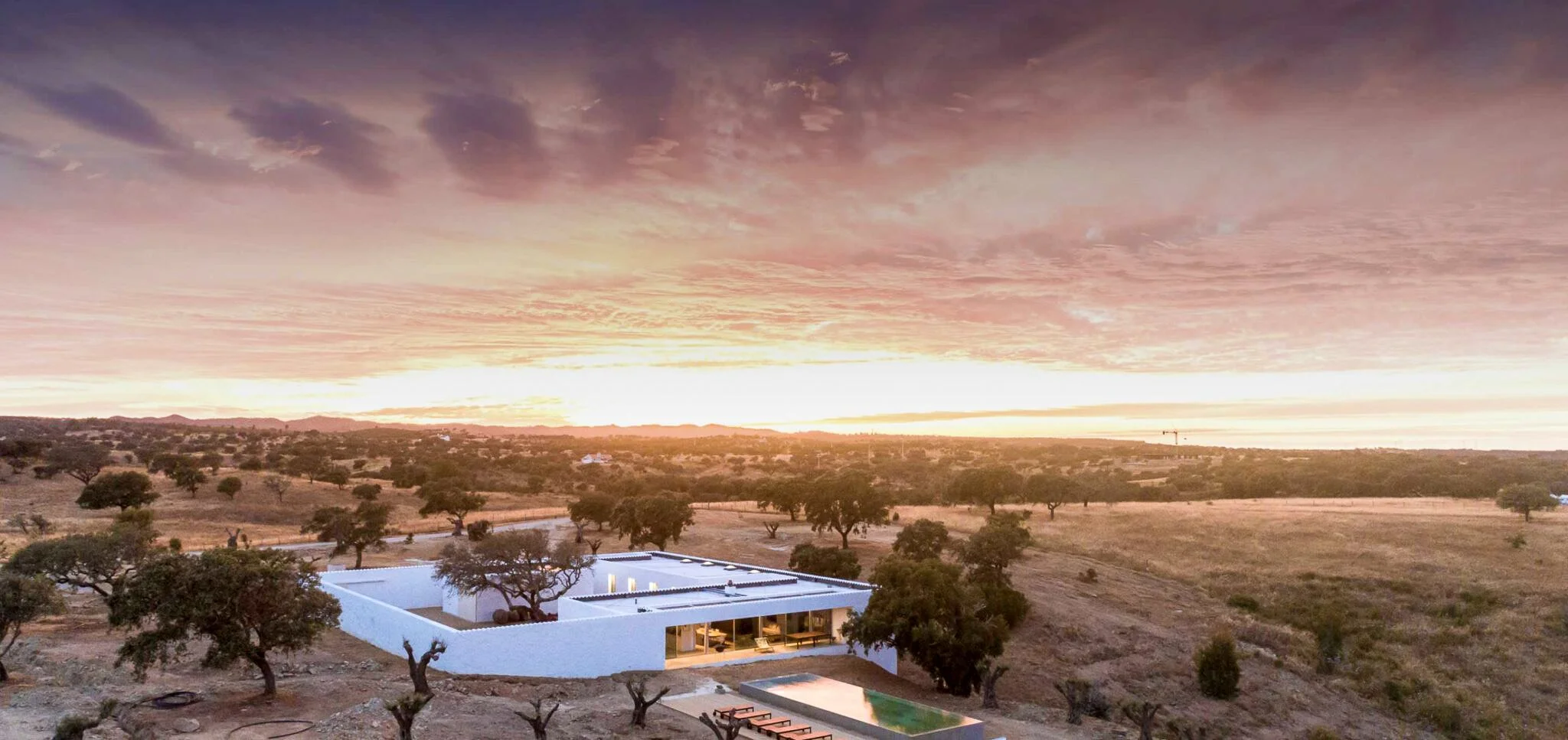 A film and photo location in Portugal from walnut films, A modern white house with a swimming pool in a desert landscape during sunset, with trees and hills in the background and a colorful sky.