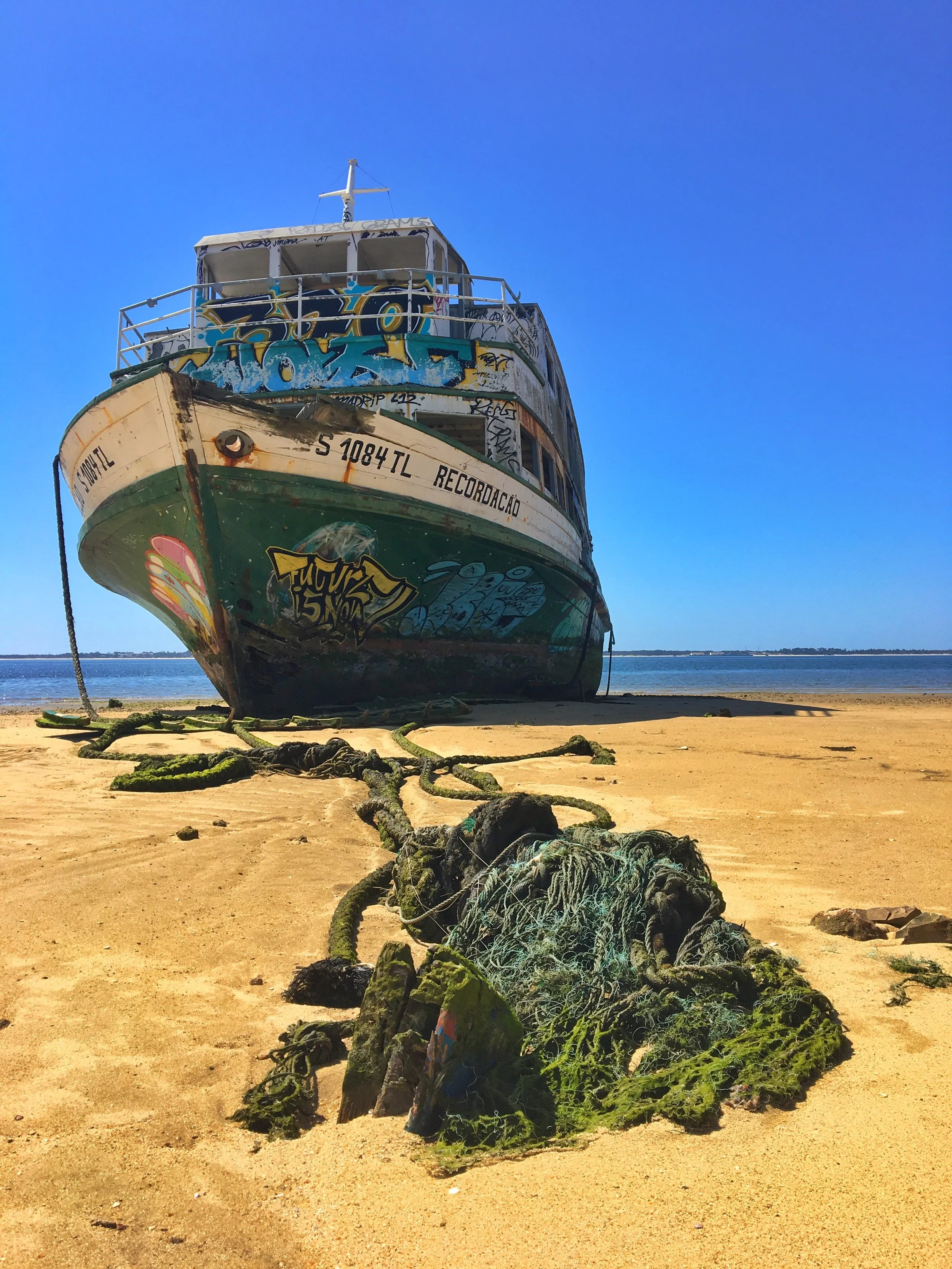 A film and photo location in Portugal from walnut films, An old, abandoned boat with graffiti on its sides, beached on sandy shore, with a large tangled rope and seaweed in the foreground, and calm water with a distant shoreline in the background.