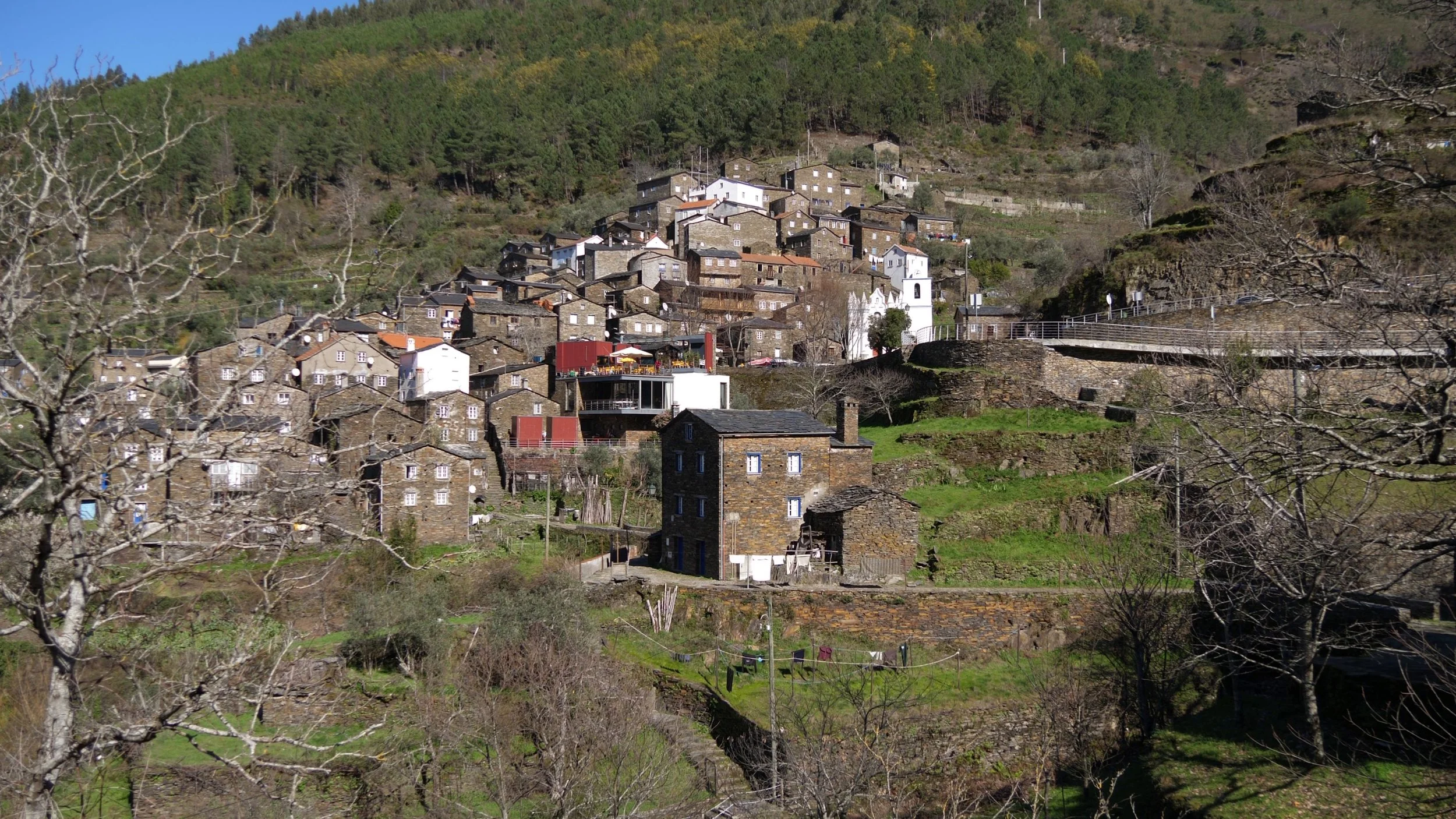 A film and photo location in Portugal from walnut films, A hillside village with stone houses and a church, surrounded by trees and greenery, under a clear blue sky.