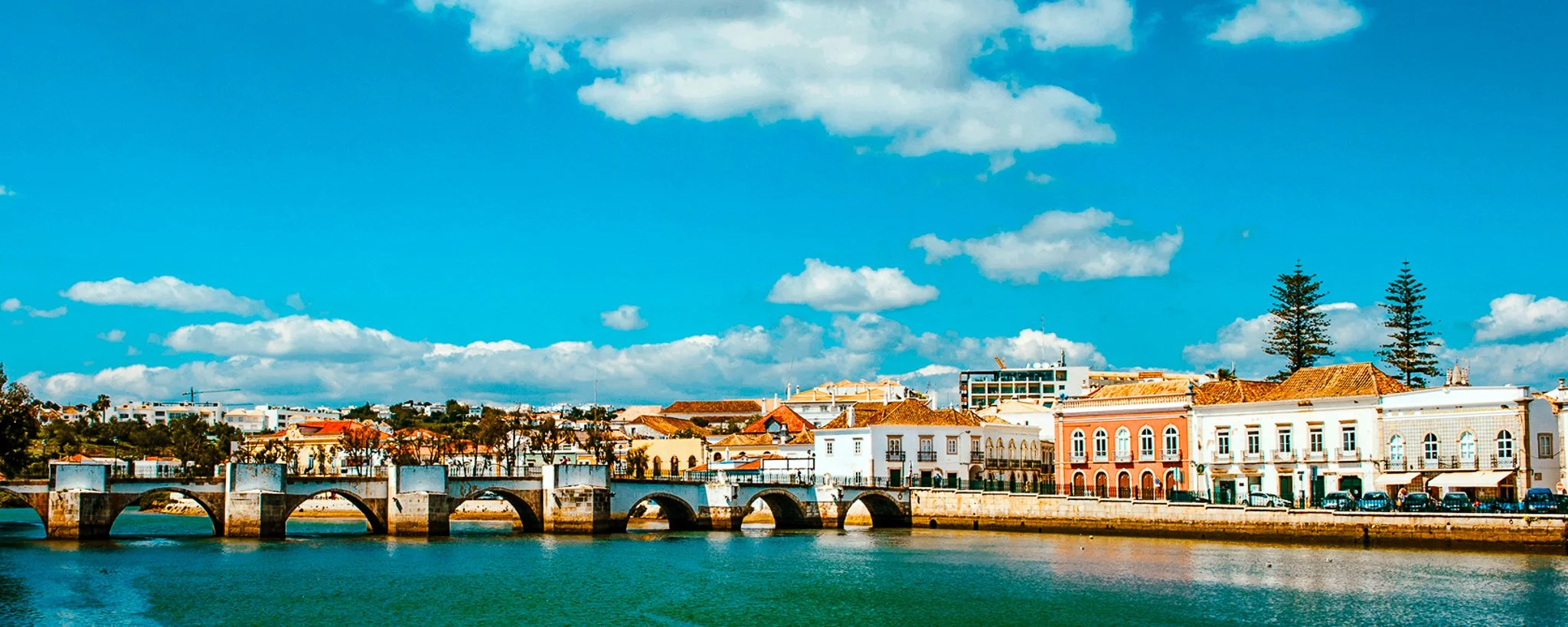 A film and photo location in Portugal from walnut films, A scenic view of a waterway with a stone bridge, colorful historic buildings along the waterfront, and tall trees under a partly cloudy blue sky.