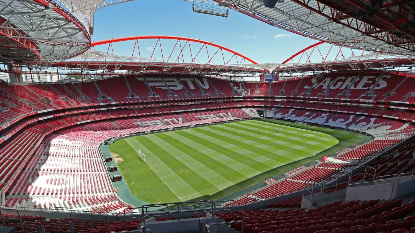 A film and photo location in Portugal from walnut films, Interior view of a large soccer stadium with red seats and green grass field, under a clear blue sky.