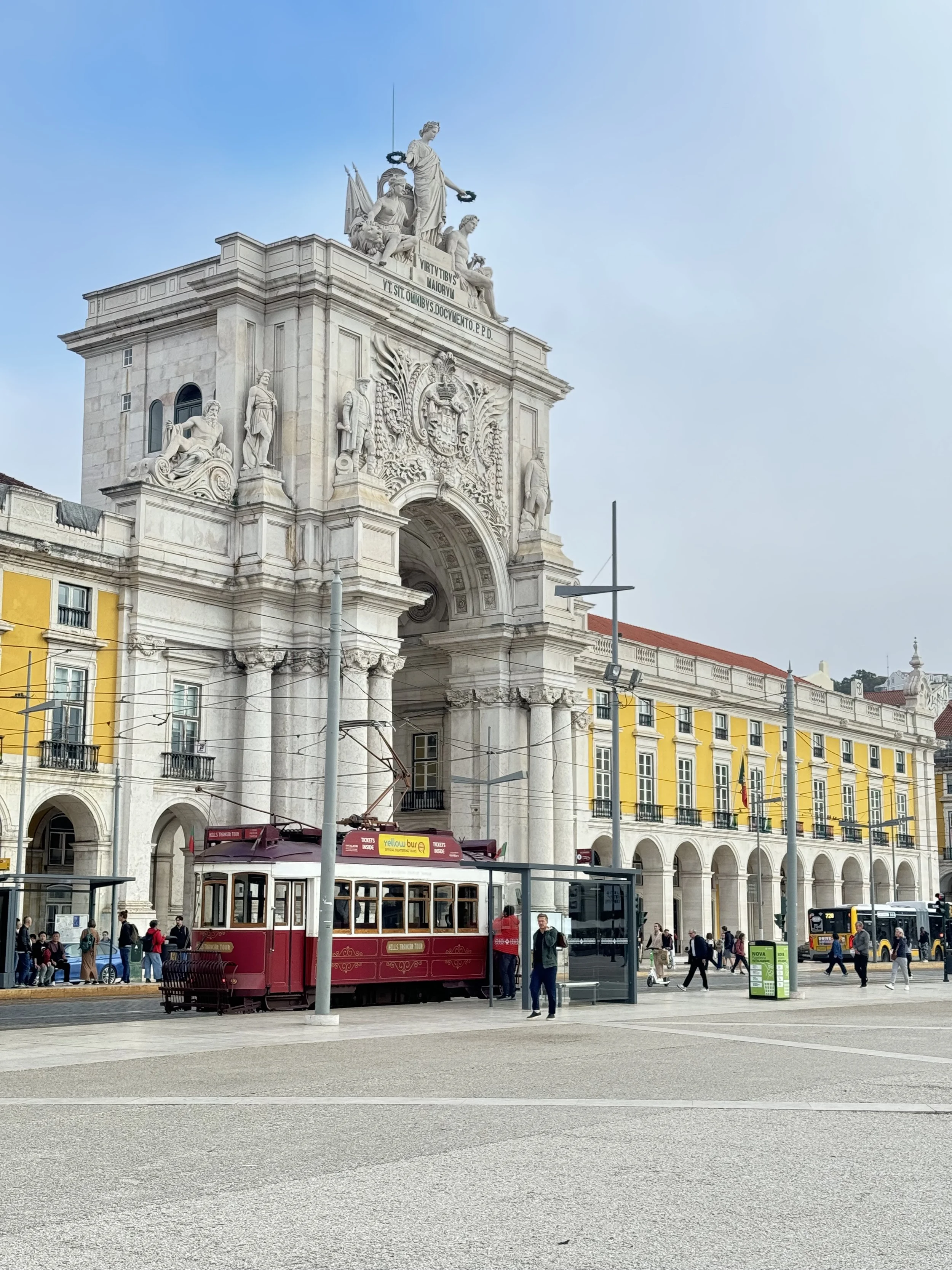 A film and photo location in Portugal from walnut films, A city square with a historic building featuring a large archway, statues, and elaborate architectural details. There is a red tram, people walking, and a trolley stop with a green sign. 