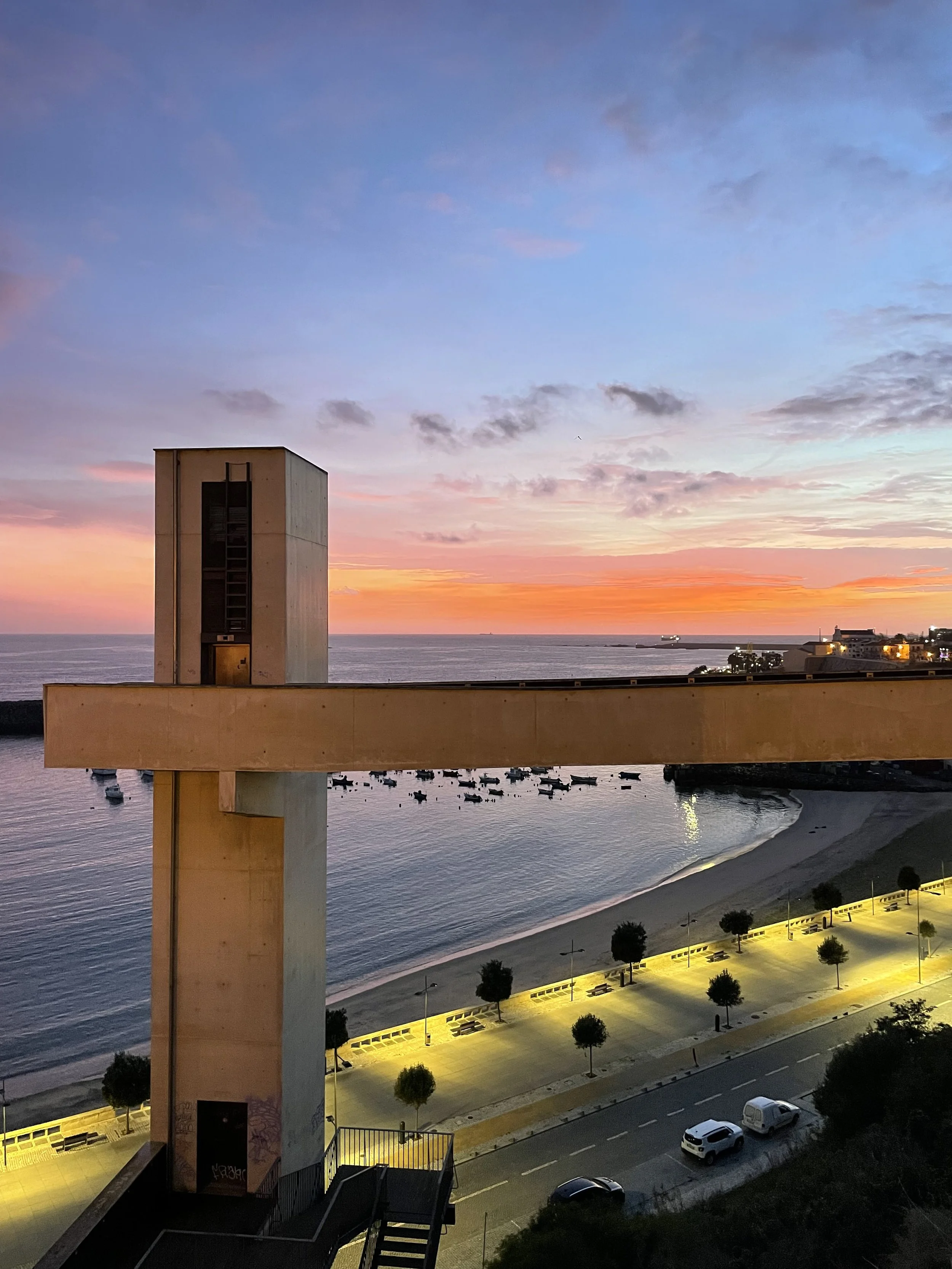 A film and photo location in Portugal from walnut films,  beachside cityscape at sunset with a large concrete building in the foreground, a parking lot with several cars, and a row of lit streetlights along a boardwalk. 