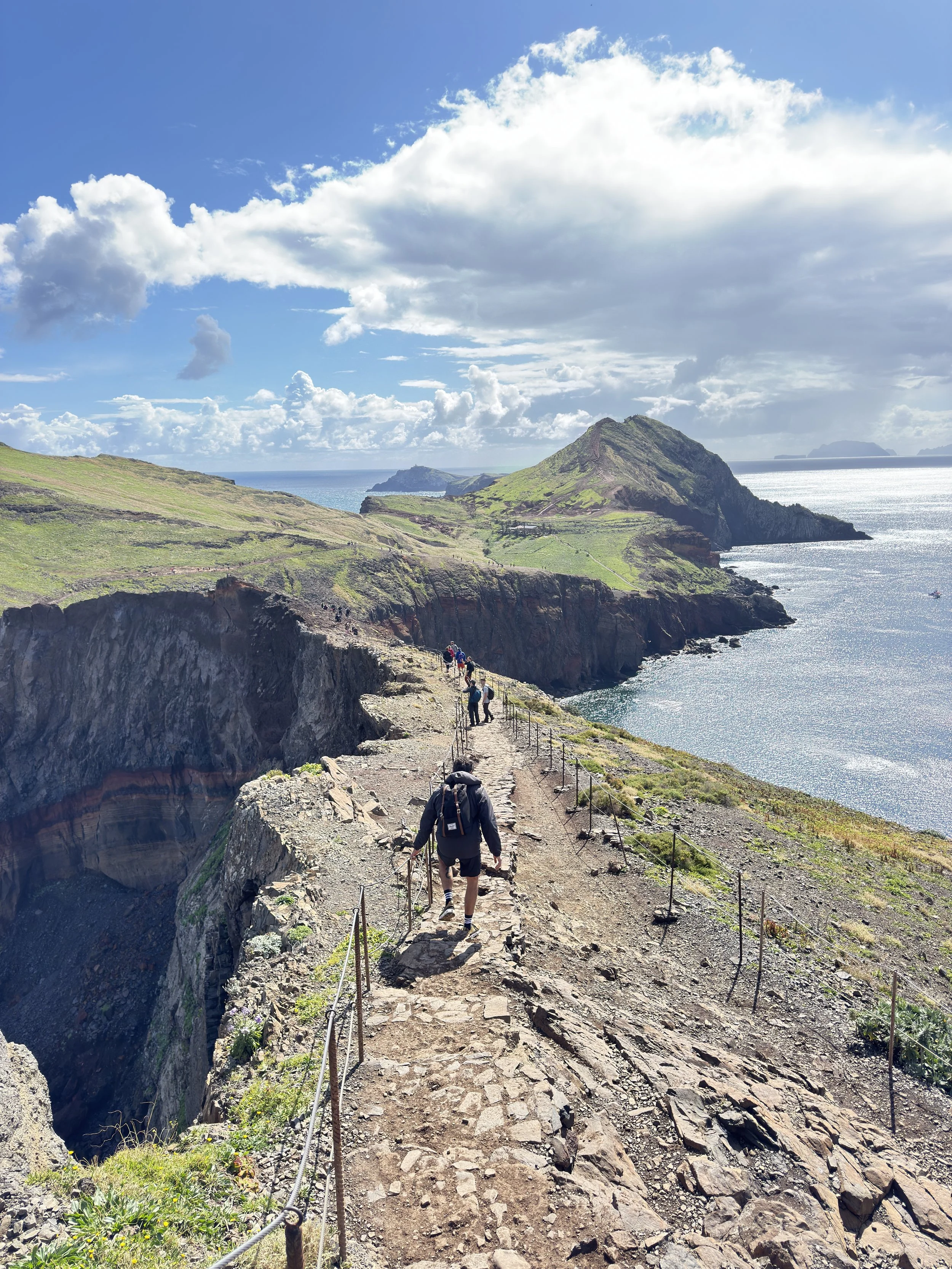 A film and photo location in Portugal from walnut films, A group of hikers walking along a rocky trail on a coastal cliff with green hills, a blue ocean, and a partly cloudy sky in the background.