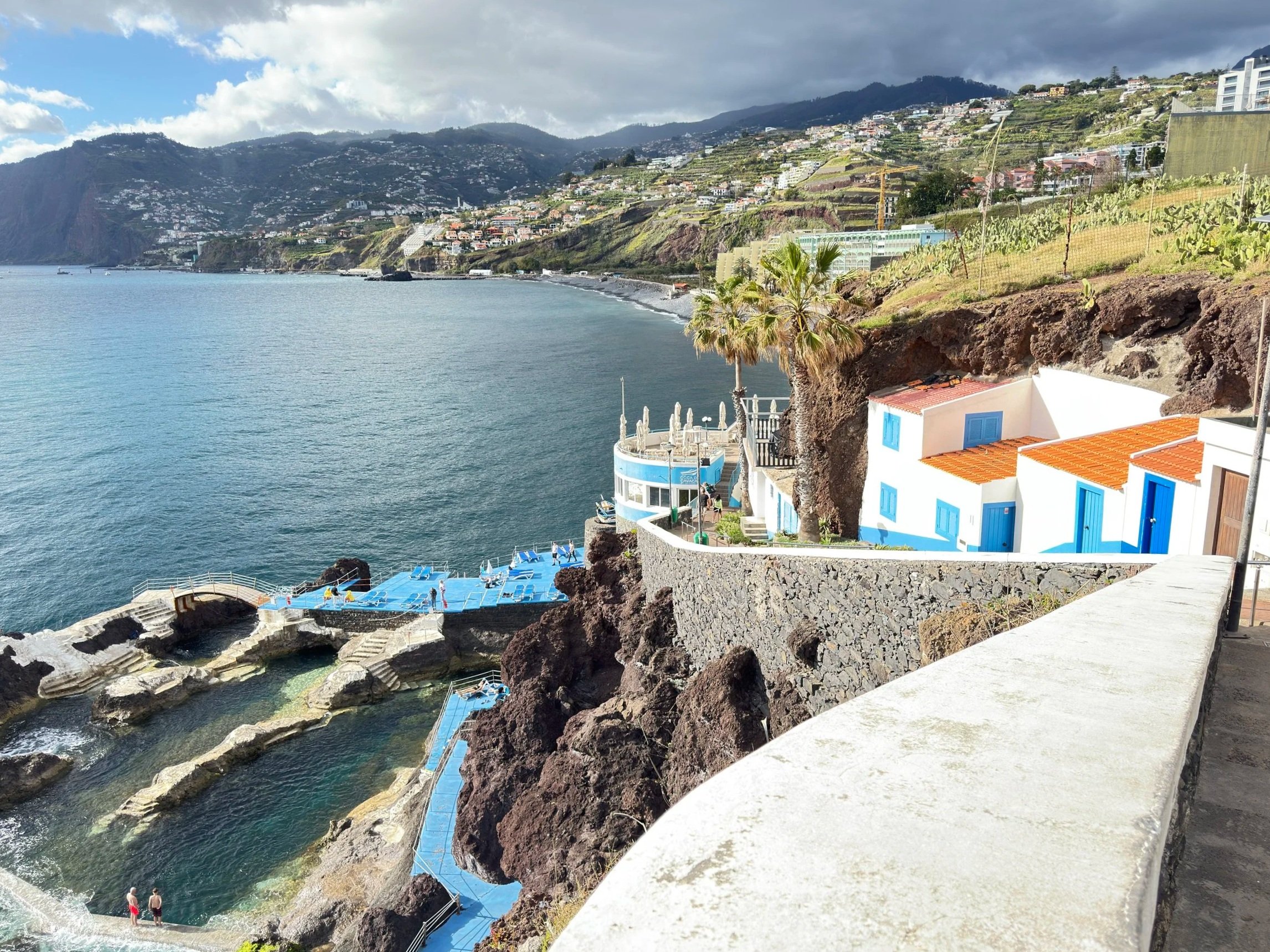 A film and photo location in Portugal from walnut films, Coastal hillside with white buildings and terraced gardens overlooking the sea, featuring a pool and two people walking along the rocky shore.