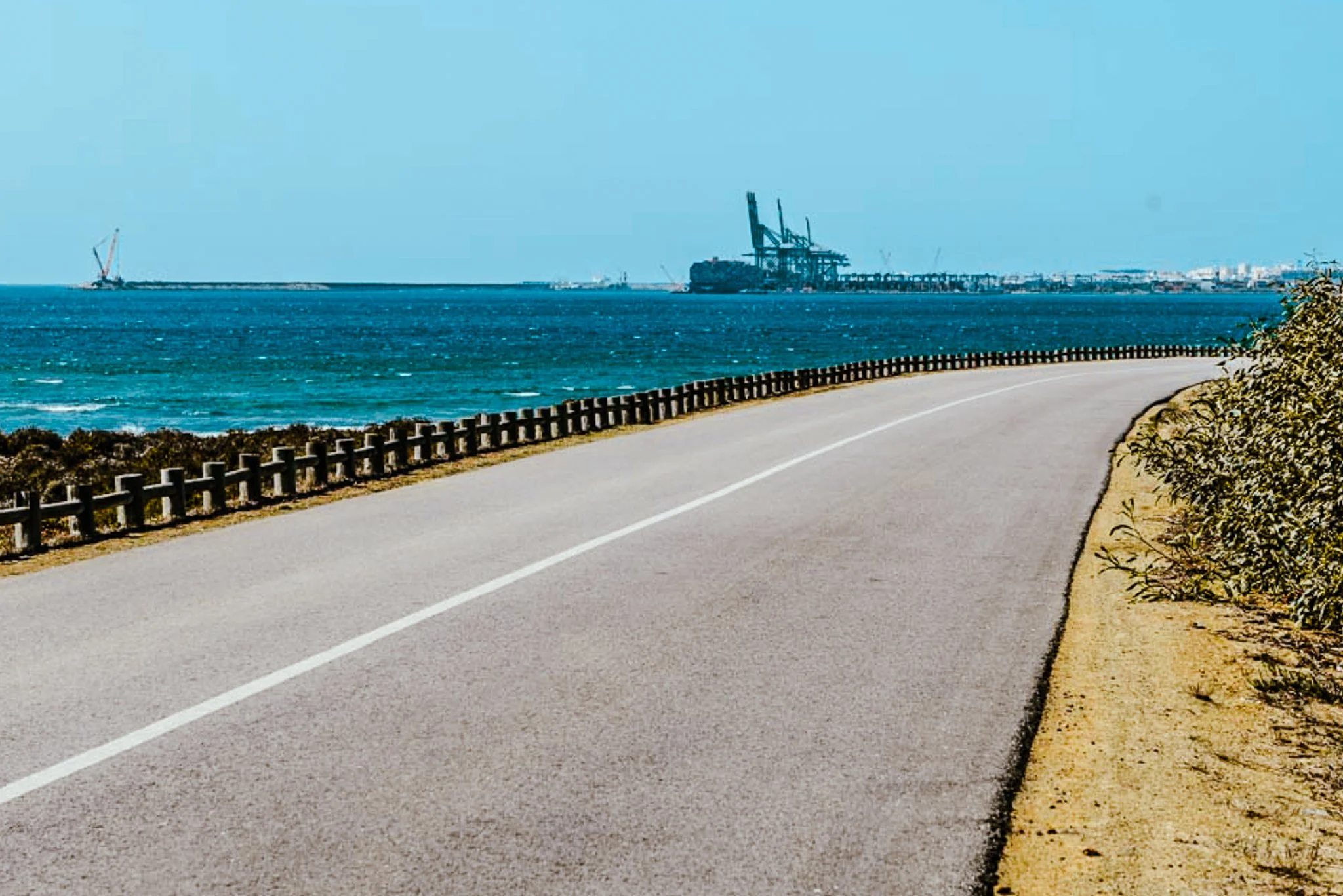 A film and photo location in Portugal from walnut films, A scenic coastal road runs alongside the ocean with a guardrail on the left and a strip of sandy land and bushes on the right. Offshore, 