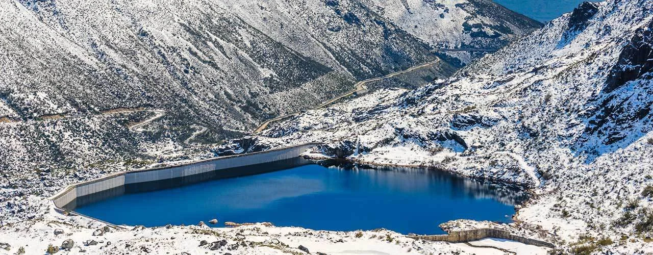 A film and photo location in Portugal from walnut films, A snow-covered mountain landscape with a winding road and a large, curved reservoir or dam in the foreground.