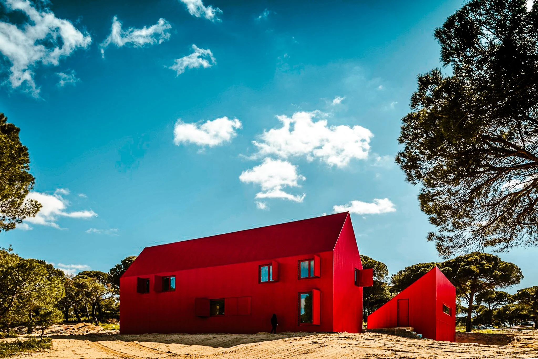 A film and photo location in Portugal from walnut films, Modern red building with sharp angular roofs under a partly cloudy blue sky, surrounded by trees and sandy ground.