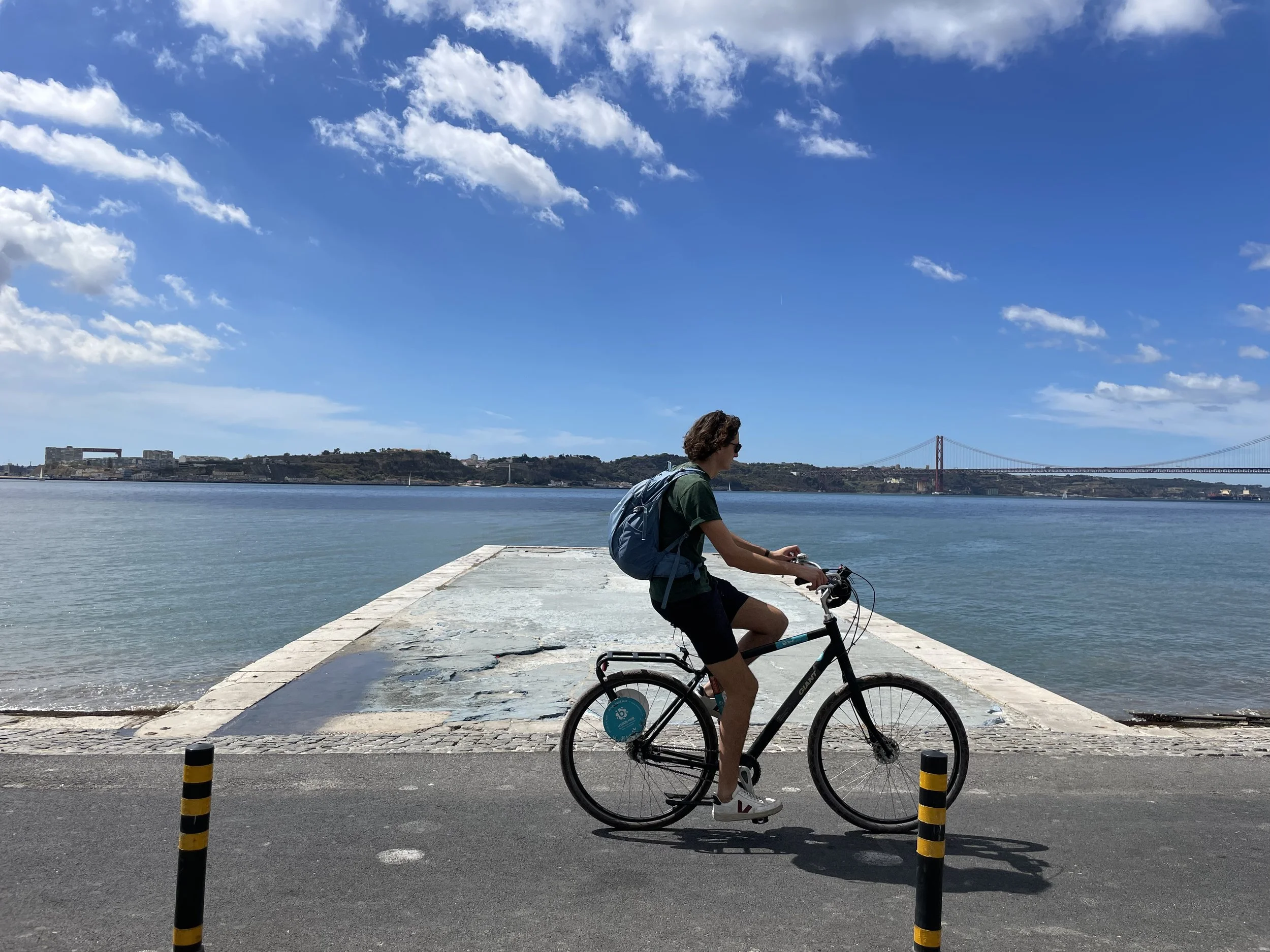 A film and photo location in Portugal from walnut films, A person riding a bicycle along a waterfront promenade with the Lisbon skyline, including the iconic 25 de Abril Bridge, in the background under a partly cloudy sky.