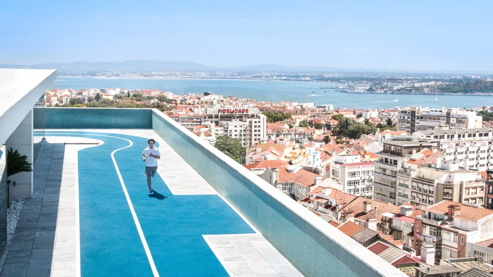 A film and photo location in Portugal from walnut films, A man jogging on a rooftop running track overlooking a city, river, and distant mountains under a clear blue sky.